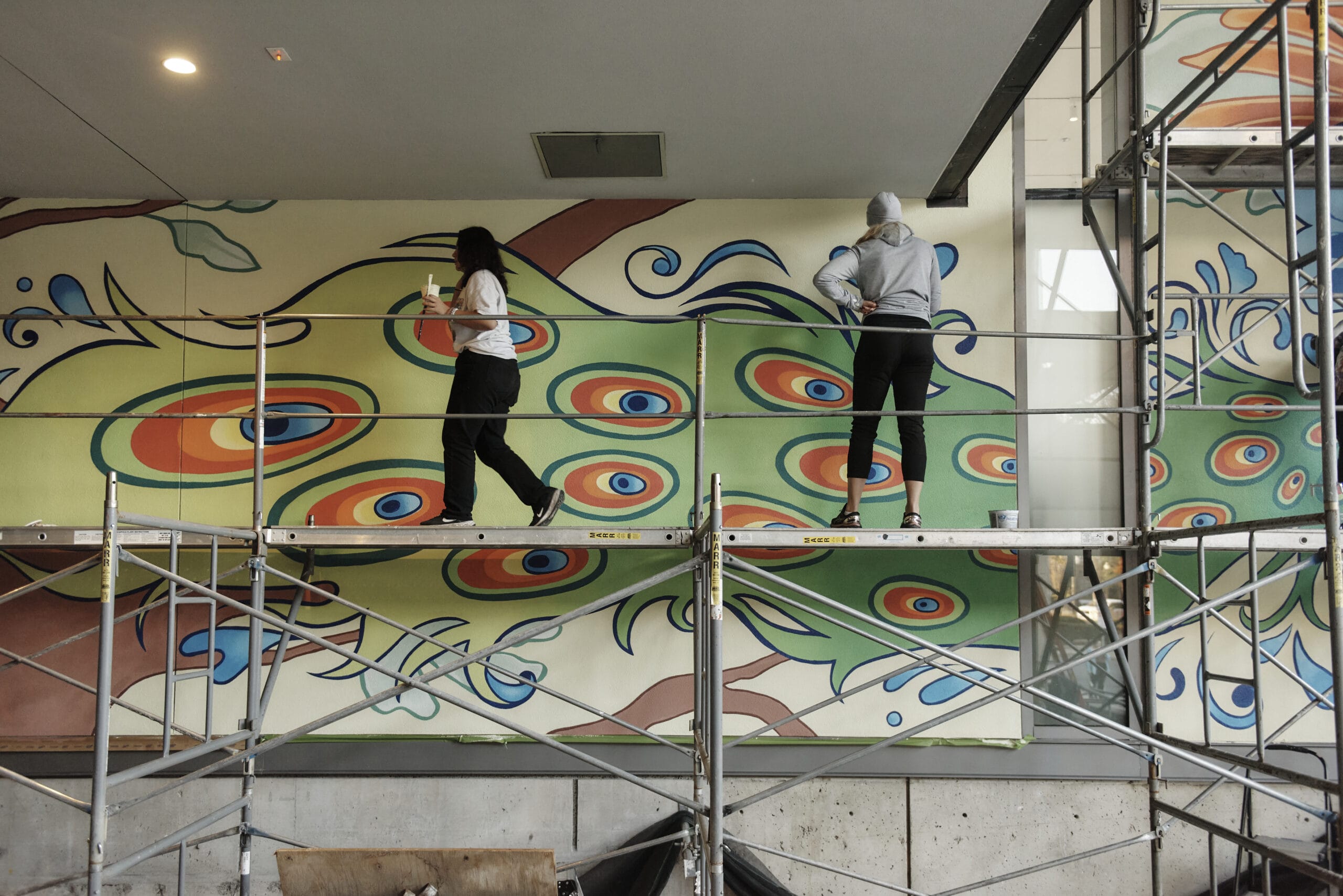 Two students stand on scaffolding working on a greek and orange mural