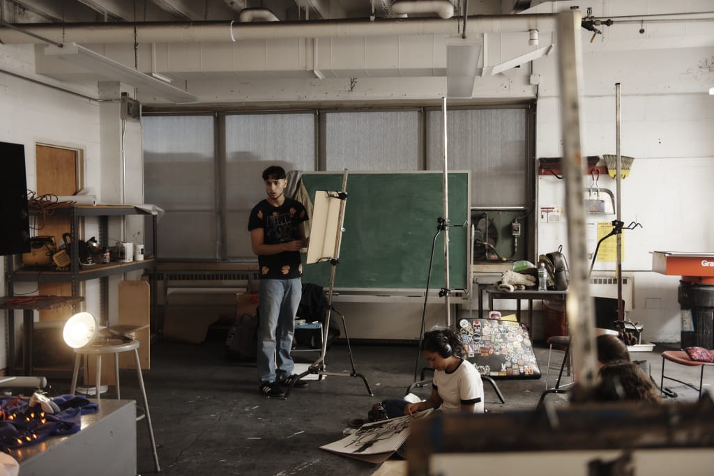 One student stands at an easel while others sit on the ground drawing in a dimly lit class with a still life.