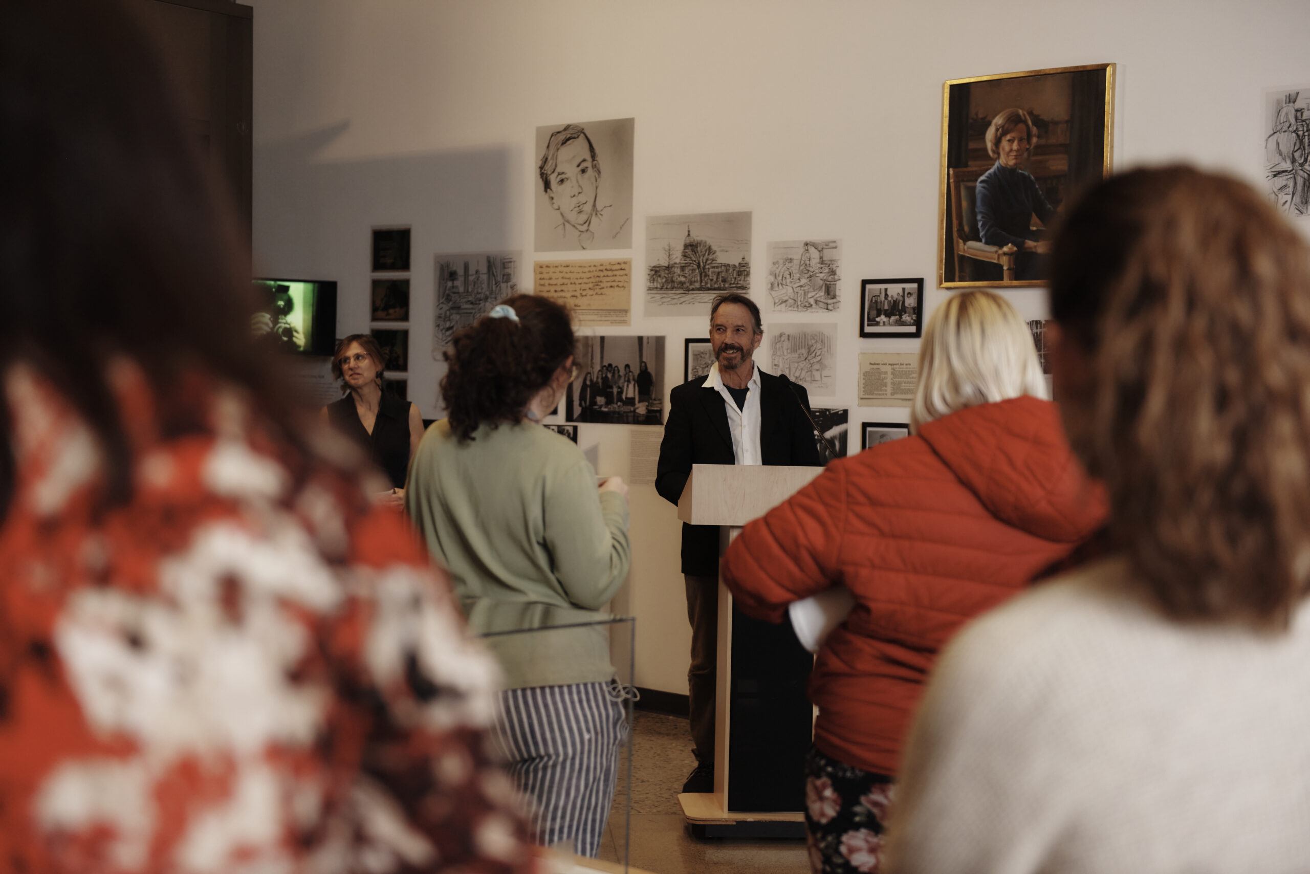 A man speaks at a podium in a room adorned with various artworks and portraits on the walls, reminiscent of a MassArt gallery. Several people stand around, listening attentively as if anticipating an auction. The atmosphere is informal yet focused.