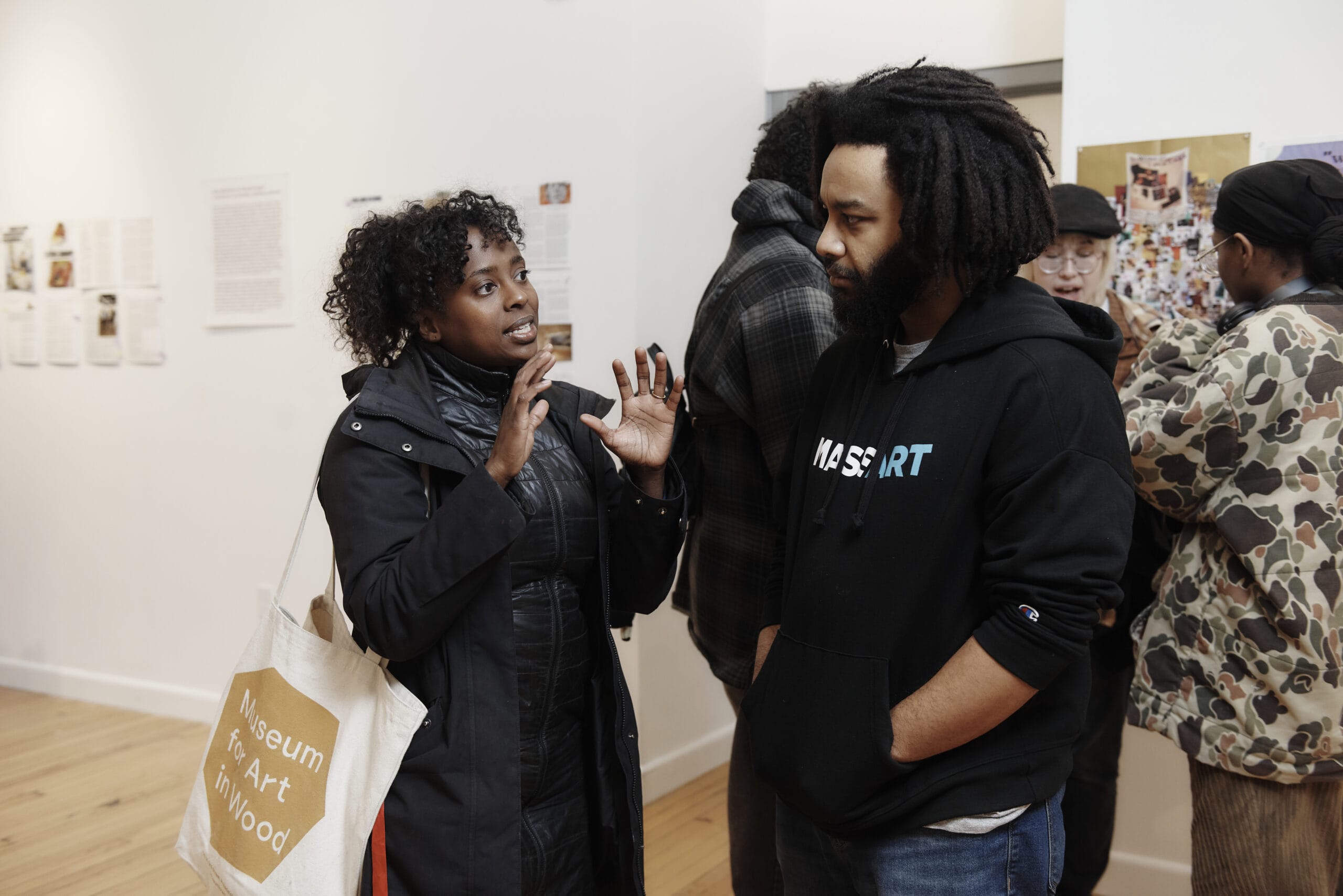 A woman with curly hair, wearing a coat and holding a tote bag, gestures animatedly while speaking to a bearded man in a hoodie. They stand in an art gallery, surrounded by others who admire the artistry on the walls.