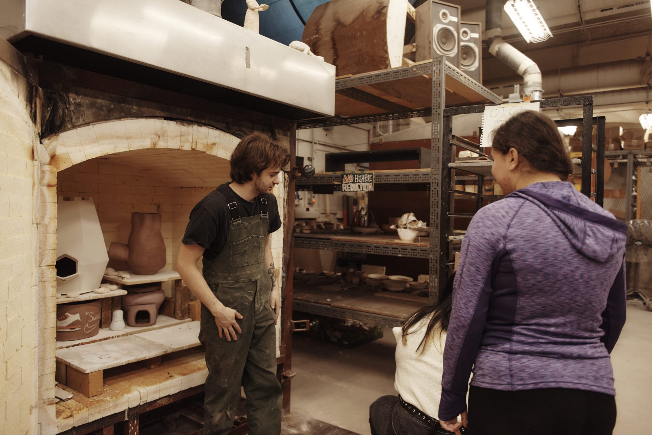 A person in overalls stands in front of an open kiln, discussing with two enrolled students in a pottery studio. Shelves filled with pottery items and equipment surround them.