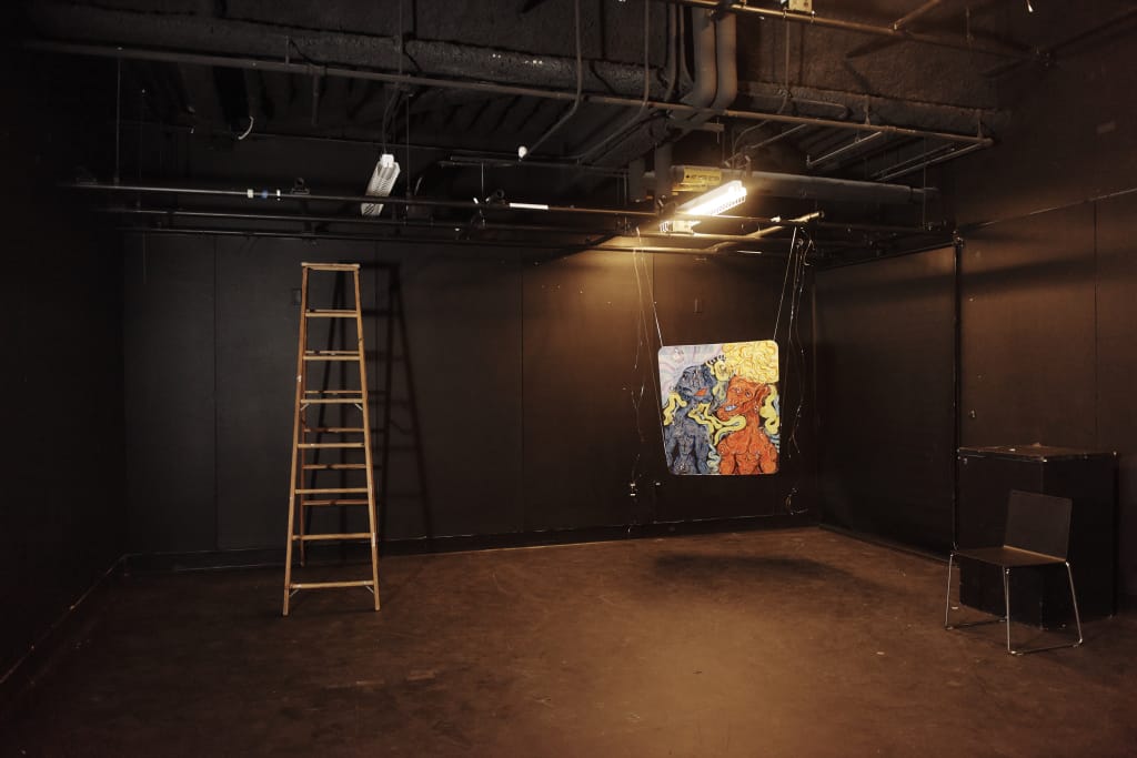 A dimly lit studio with black walls and ceiling features a wooden ladder, a colorful abstract painting on the back wall, and a black chair. Overhead, pipes and lights are visible. The space has a minimalist, industrial feel, making efficient use of resources for an artistic atmosphere.