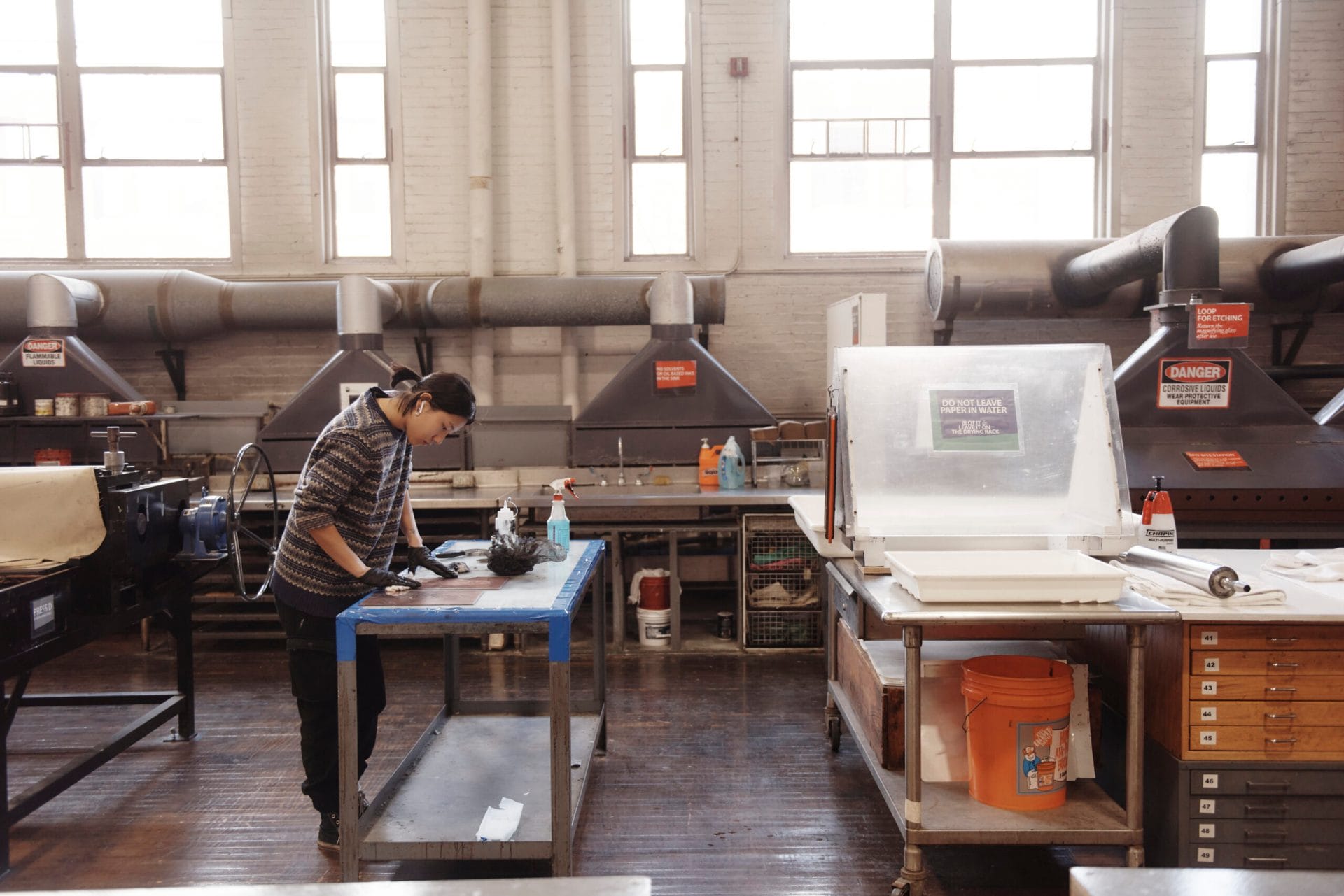 A person stands at a workbench in an industrial workshop with large windows. They are focused on a task, surrounded by equipment and tools, including large ventilation units and storage cabinets on a wooden floor.