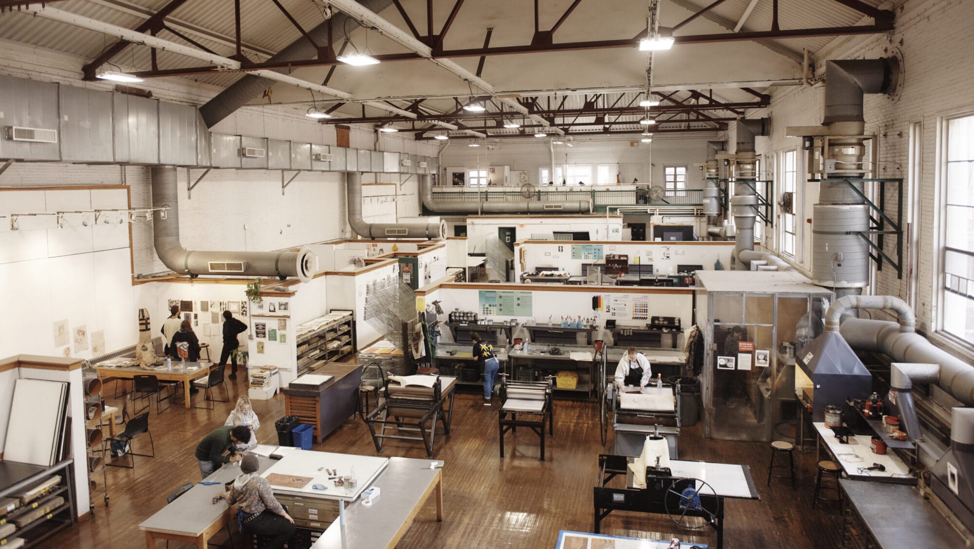 In a spacious art studio with high ceilings and large windows, artists work at various tables surrounded by art supplies, tools, and prints. The room has an industrial feel with exposed beams and visible ductwork. Many are enrolled in certificate programs to refine their craft.