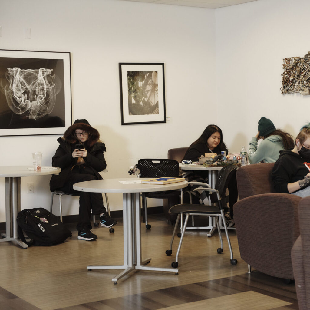 Students sitting at tables and in upholstered chairs in a white walled room with artwork