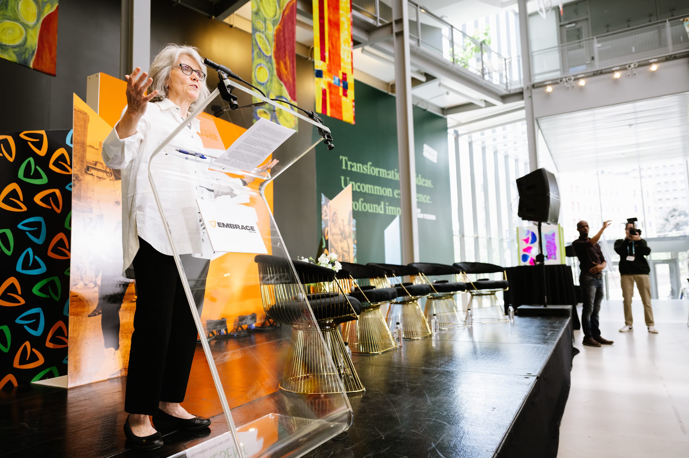 Photo behind two women speaking on a stage with President Grant in the front row watching.