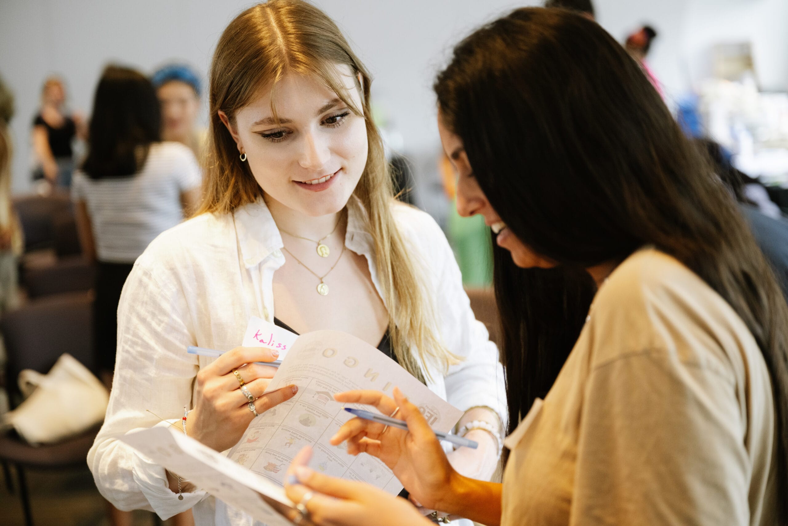 A blonde student in a white shirt smiles and talks to another student looking down at a piece of paper.