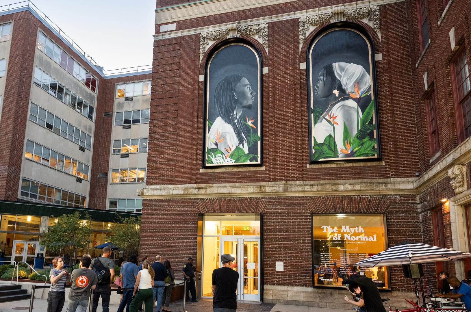 People gather outside a brick building featuring two large murals. Each mural shows an individual in profile surrounded by flowers—a scene reminiscent of MassArts creativity. Nearby, theres signage with text partially visible and a small canopy with chairs and tables, enhancing the Art Museum vibes.