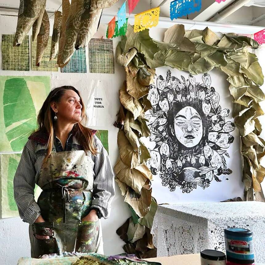 Artist in a paint-splattered apron stands in a studio with artworks and colorful decorations.