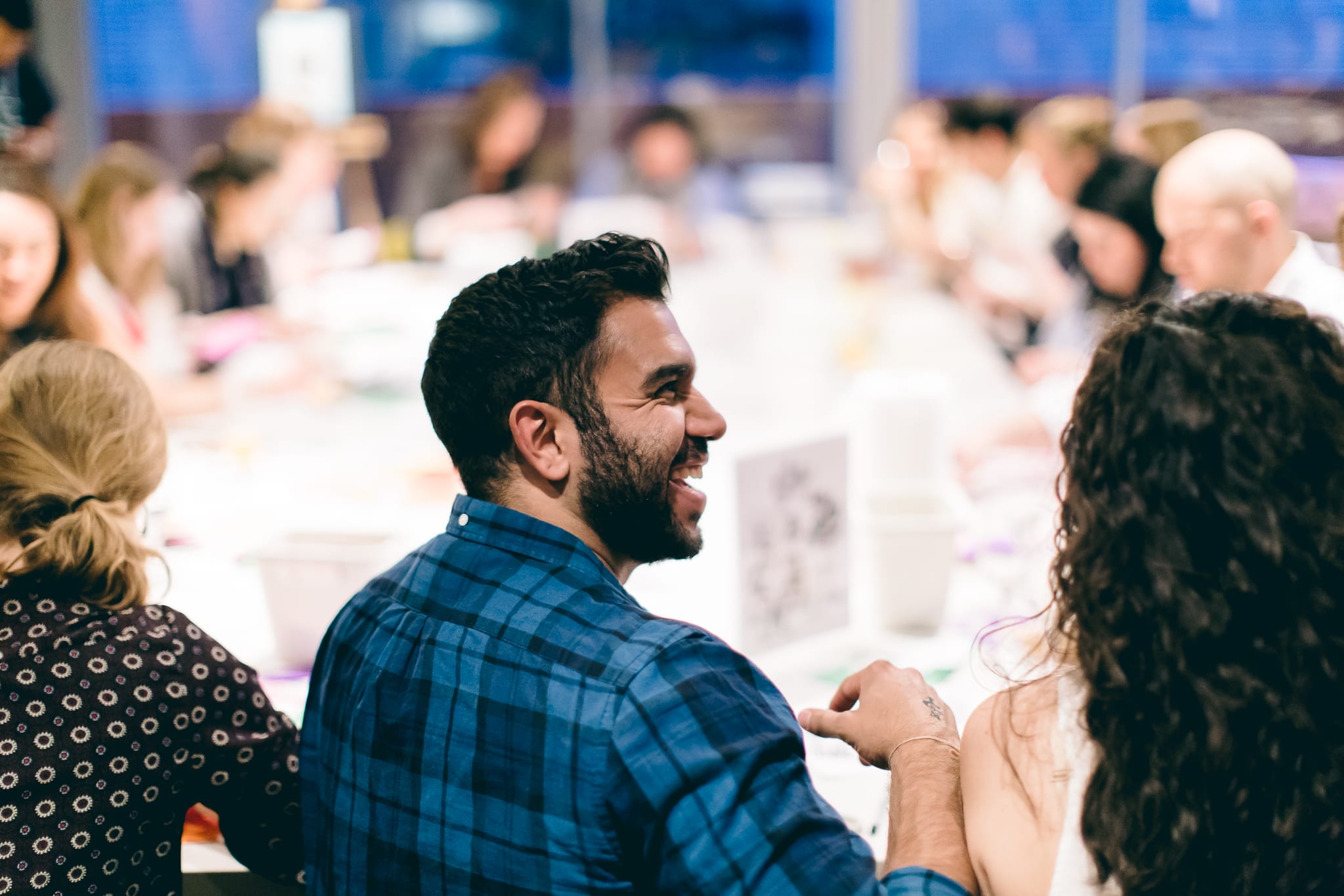 A man in a blue checkered shirt is smiling at a table crowded with people discussing college costs amidst lively activities. The background is slightly blurred, capturing a vibrant and social atmosphere.
