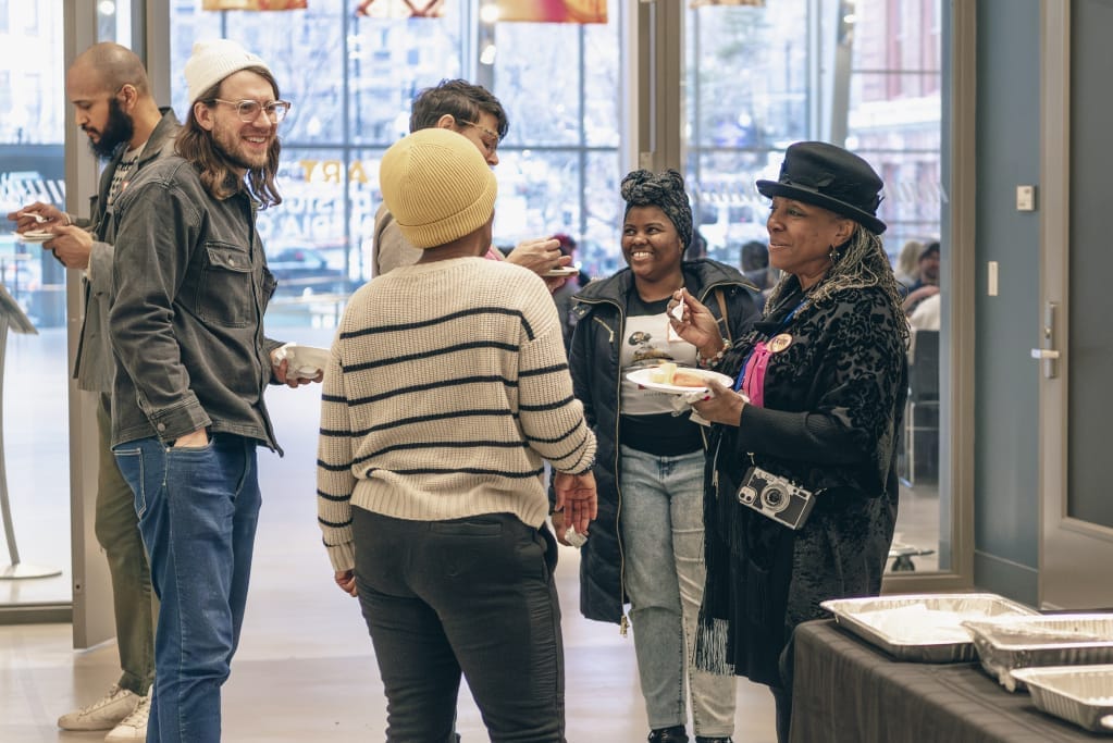 A group of people stand and chat in a room with large windows, embodying the spirit of community. Casually dressed, some hold plates of food. The background shows tables with trays, suggesting partnerships in event planning. The atmosphere is lively and social, reflecting a shared artistic flair.