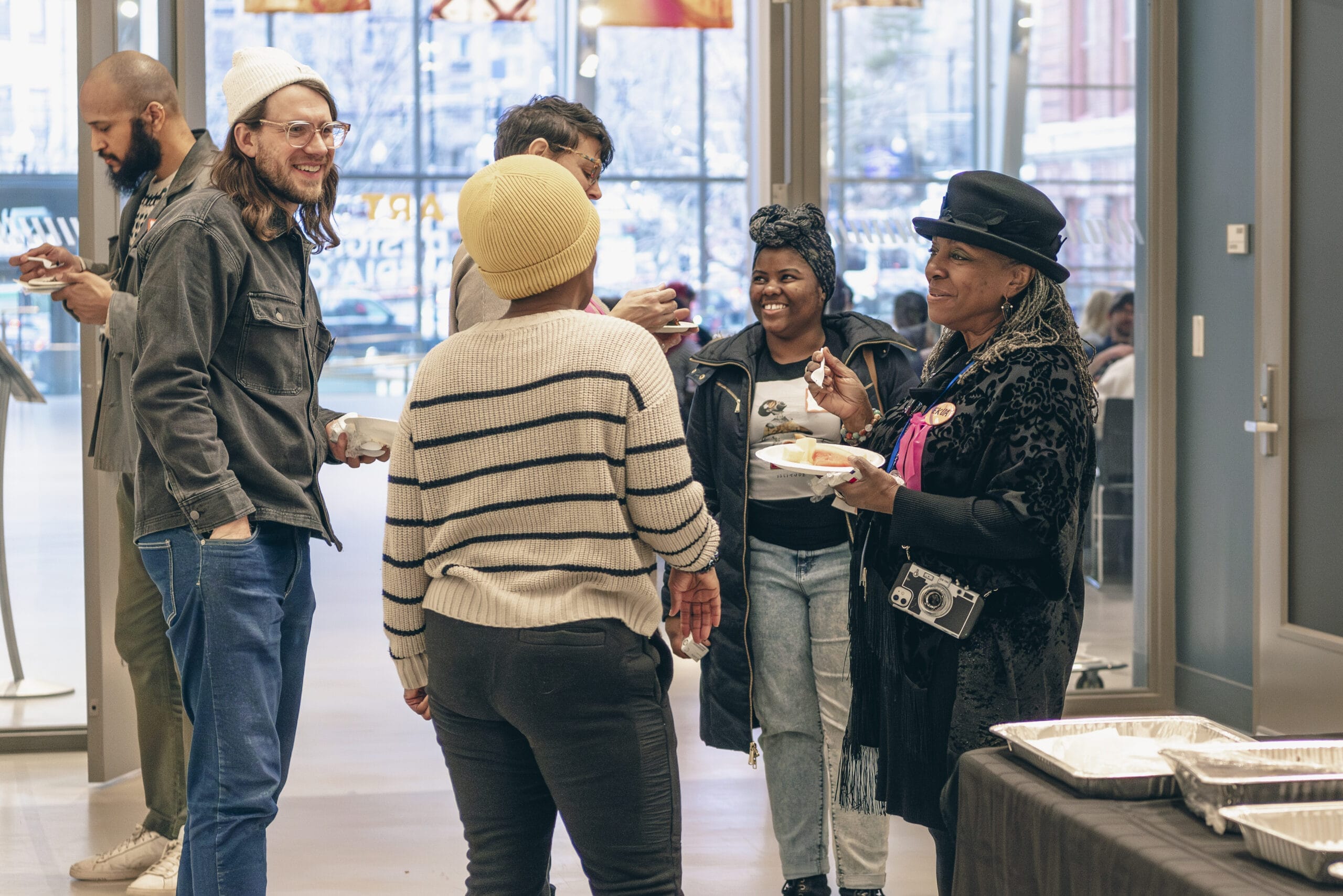 A group of people stand and chat in a room with large windows, embodying the spirit of community. Casually dressed, some hold plates of food. The background shows tables with trays, suggesting partnerships in event planning. The atmosphere is lively and social, reflecting a shared artistic flair.