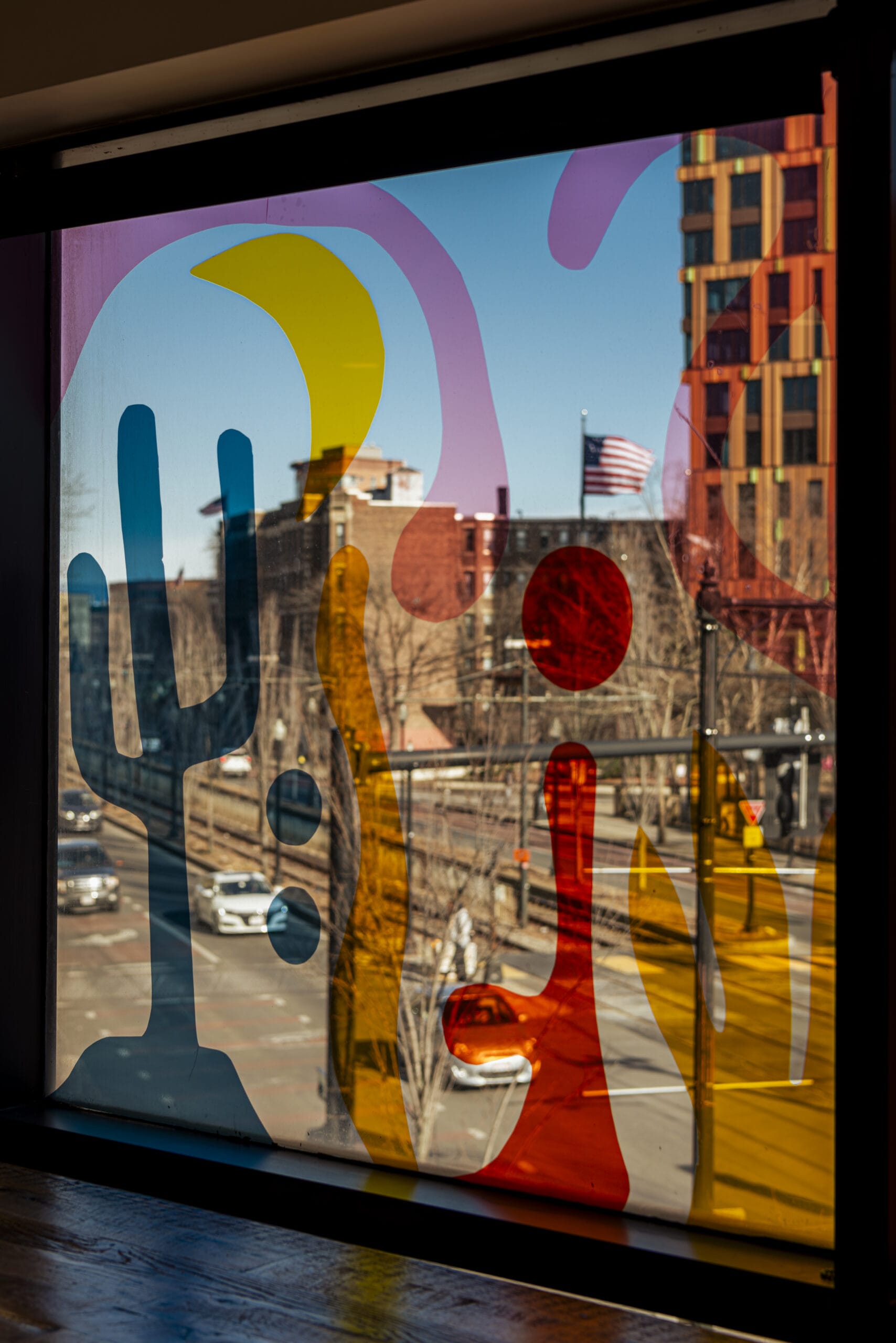 Abstract shapes in red, yellow, and purple adorn a window at Aspire@MassArt, revealing a vibrant city street scene. Through the transparent patterns, cars zoom by trees and buildings beneath a waving American flag.