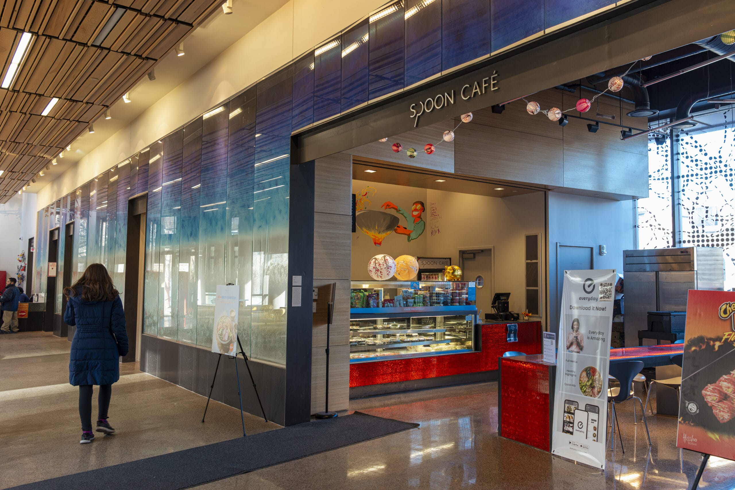 A person in a blue coat walks toward Sjoon Café, which features a modern design with a summer-inspired blue and red color scheme. The café showcases a selection of pastries and menu items inside a glass display case. Bright, decorative lights hang overhead.