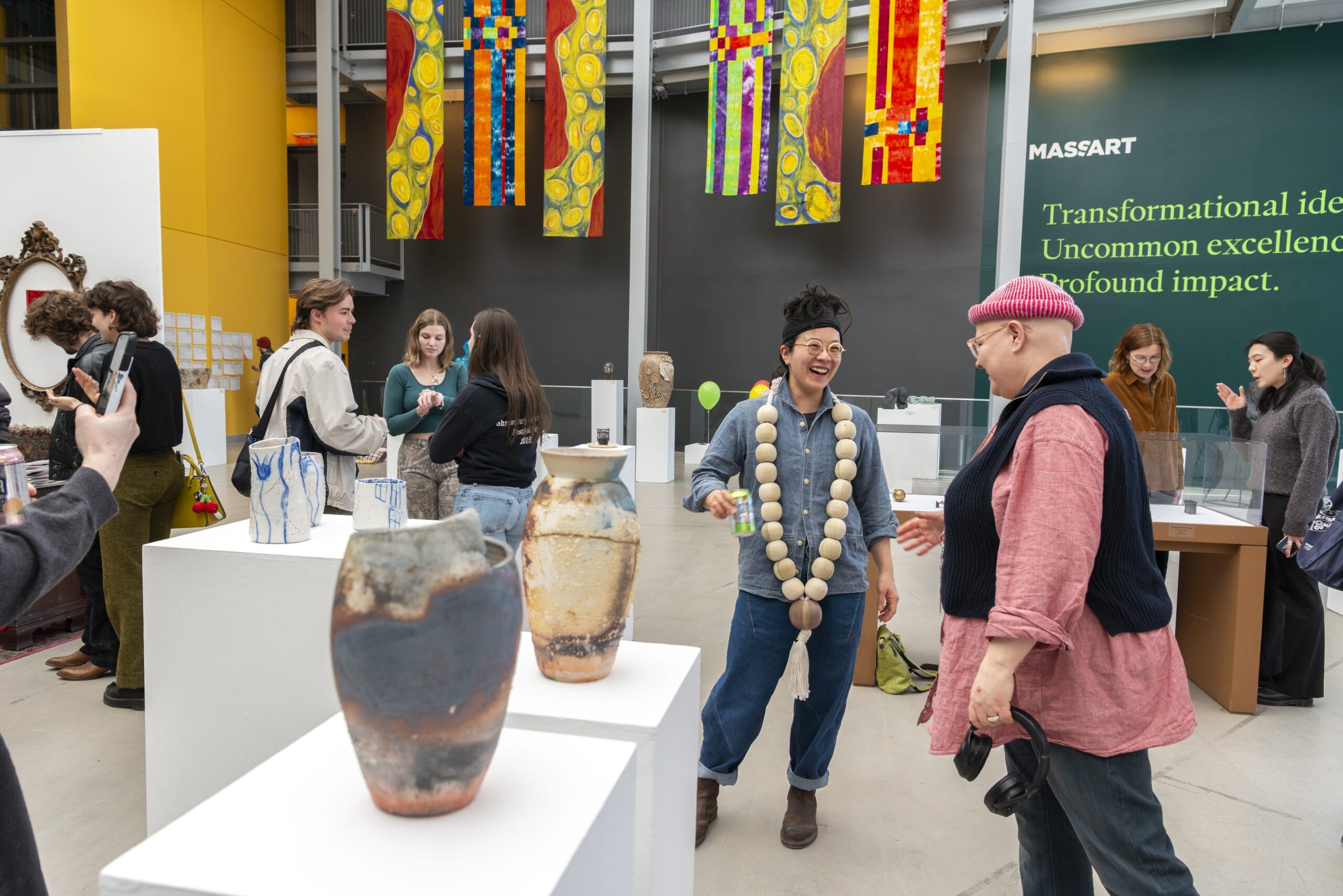 At the MassArt exhibit, people interact amid ceramic vases on pedestals. Two individuals share a laugh under vibrant banners, while others admire the artwork. The gallery buzzes with creativity and community spirit, embodying the essence of artistic work and expression.