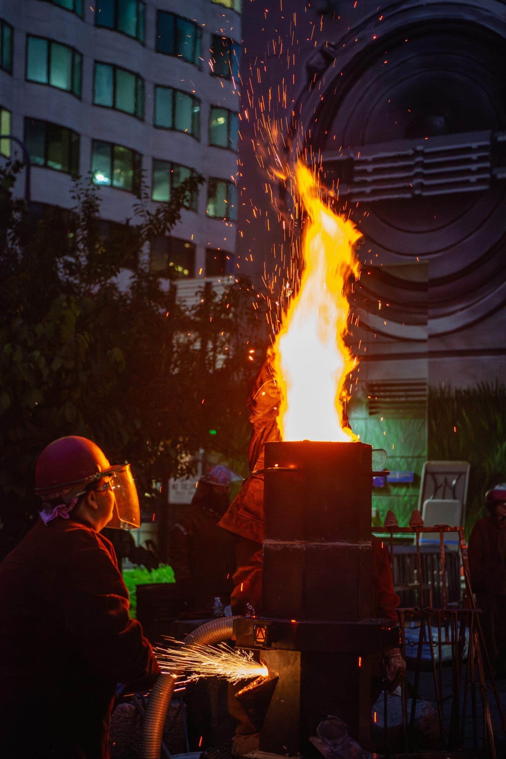 A person wearing protective gear operates a tall furnace with bright flames shooting upwards. The scene is set outdoors in an urban area, with buildings visible in the background.