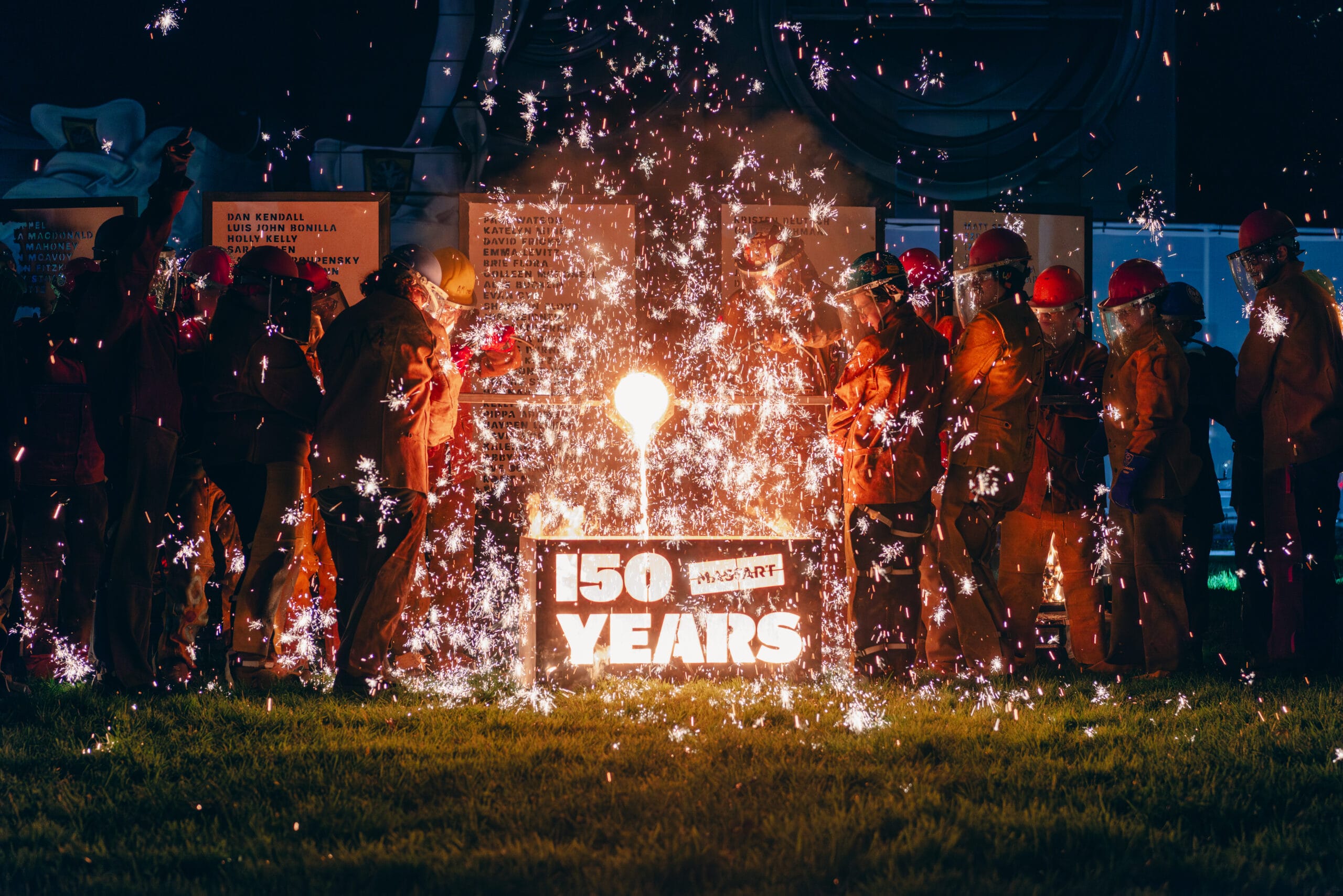 MassArt 150 Years logo is cut out of a metal box and iron corps alumni pour molten iron into sending sparks flying in the air.