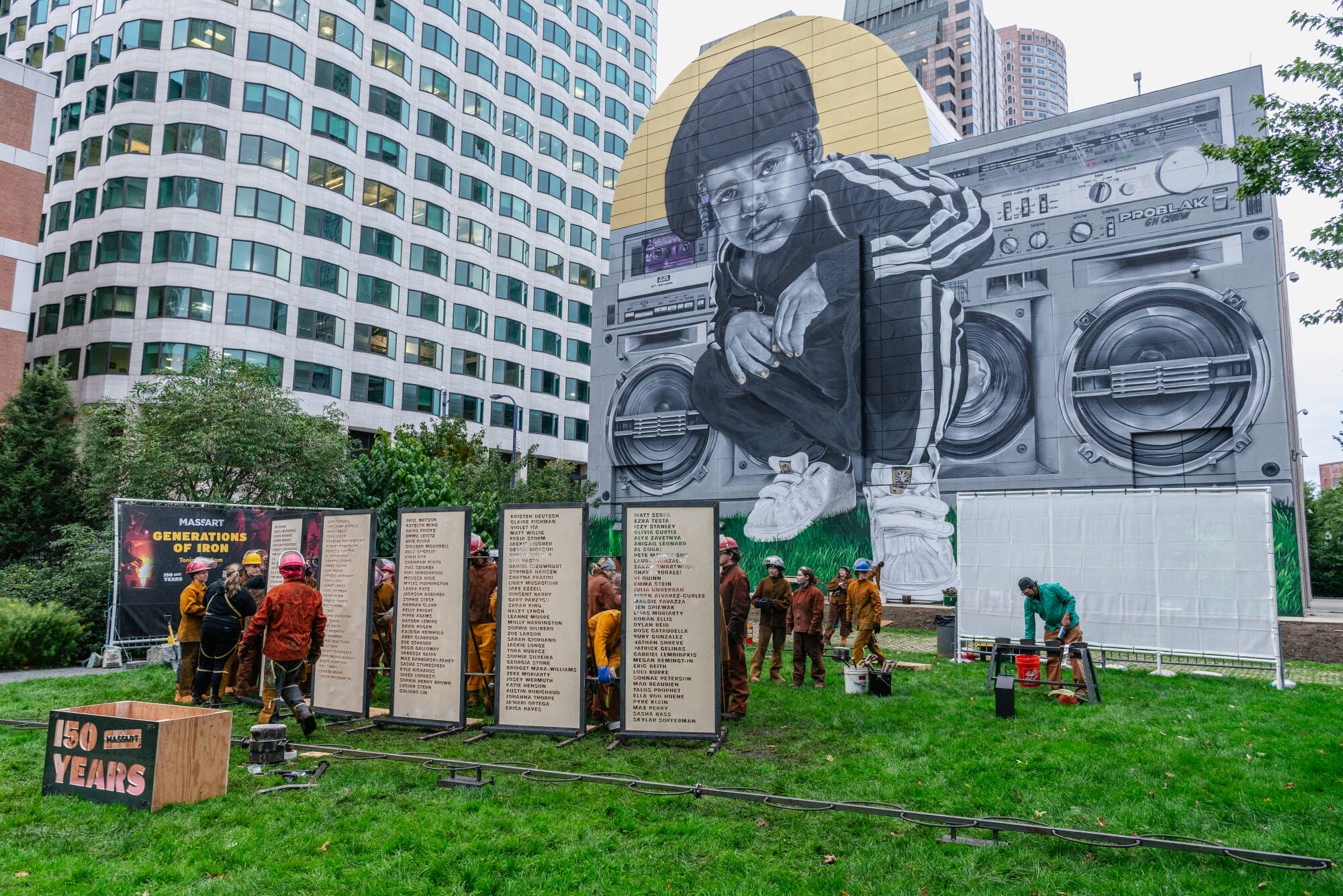 A group of people work on a Boston park project beneath a large mural of a child in urban attire sitting atop a giant boombox. The surrounding boards display information, and a 150 Years sign is visible in the foreground, encapsulating city life amidst towering buildings.
