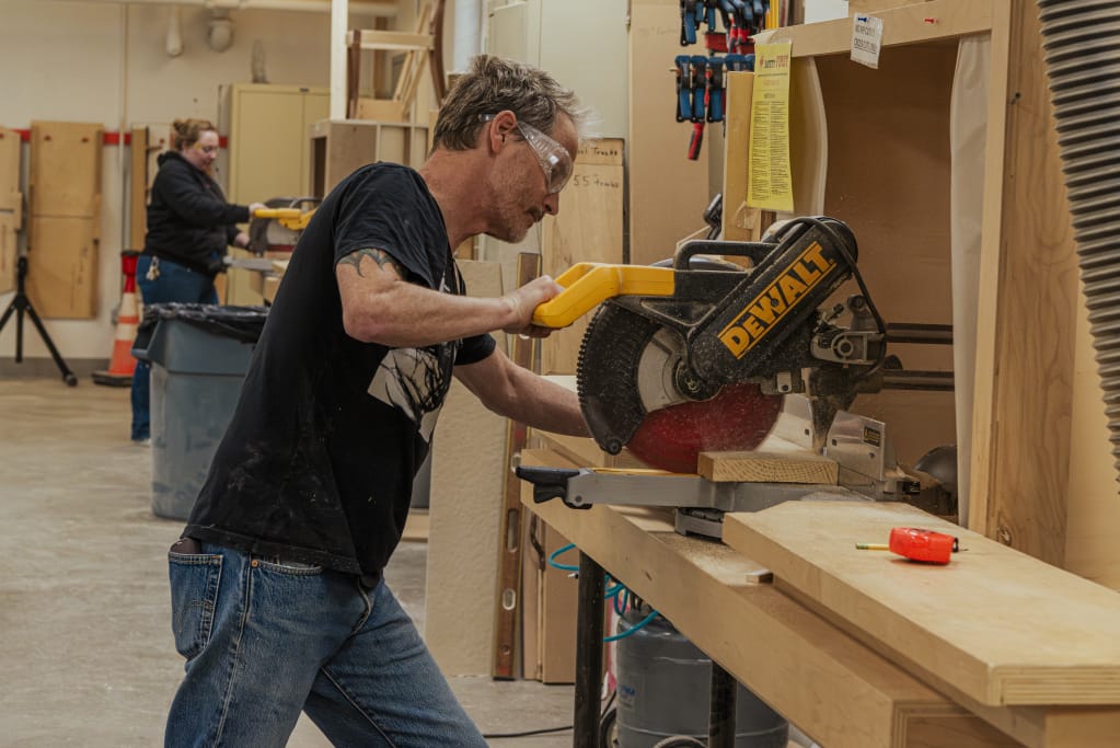 A person wearing safety goggles operates a Dewalt miter saw, cutting a wooden board in a workshop. Another person is visible in the background, engaged in woodworking tasks as part of their professional education journey. The workspace is organized with tools and materials.