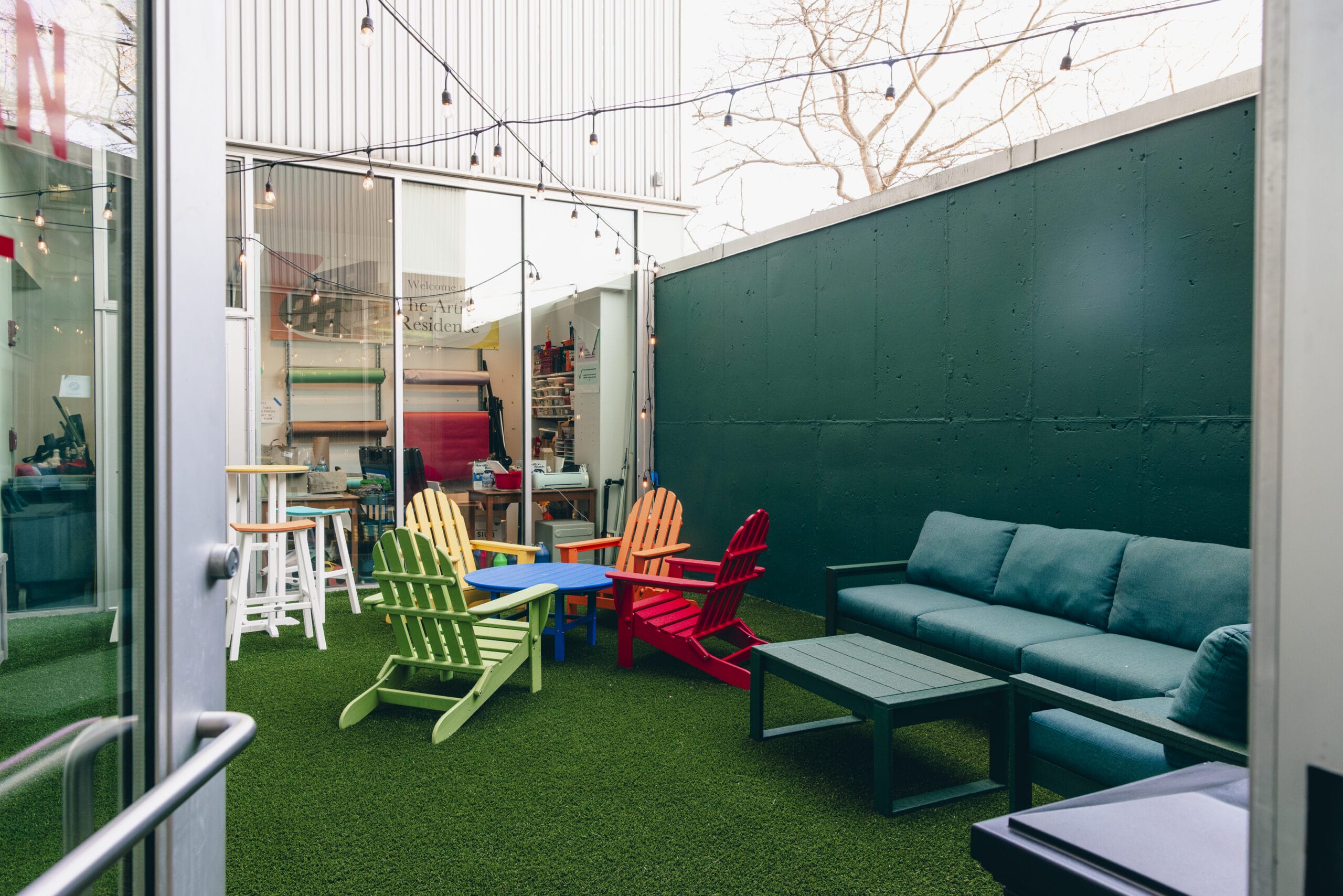 The outdoor patio, a summer housing oasis, features colorful chairs and tables on plush artificial grass. A teal sofa and wooden tables lean against a high, dark green wall. Overhead, string lights twinkle while a door on the left invites you to explore this vibrant escape.