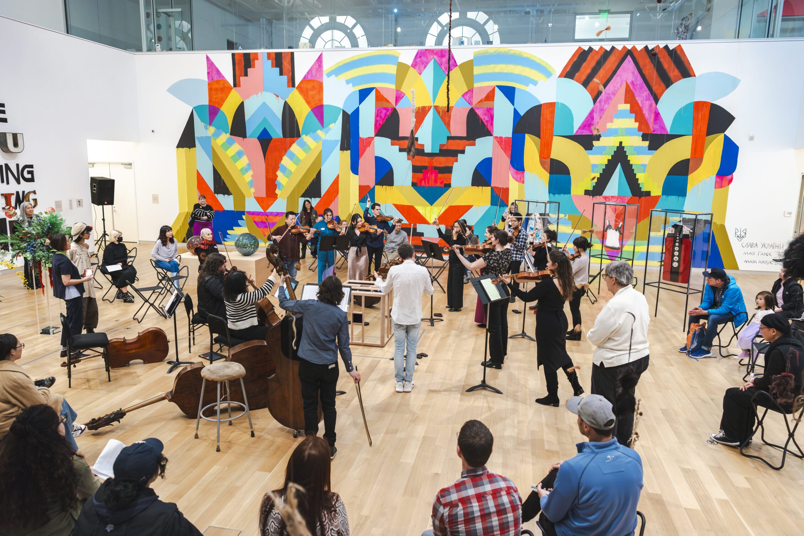 A string orchestra stands performing in the middle of an art gallery with a bright colorful artwork installed on the wall behind them.