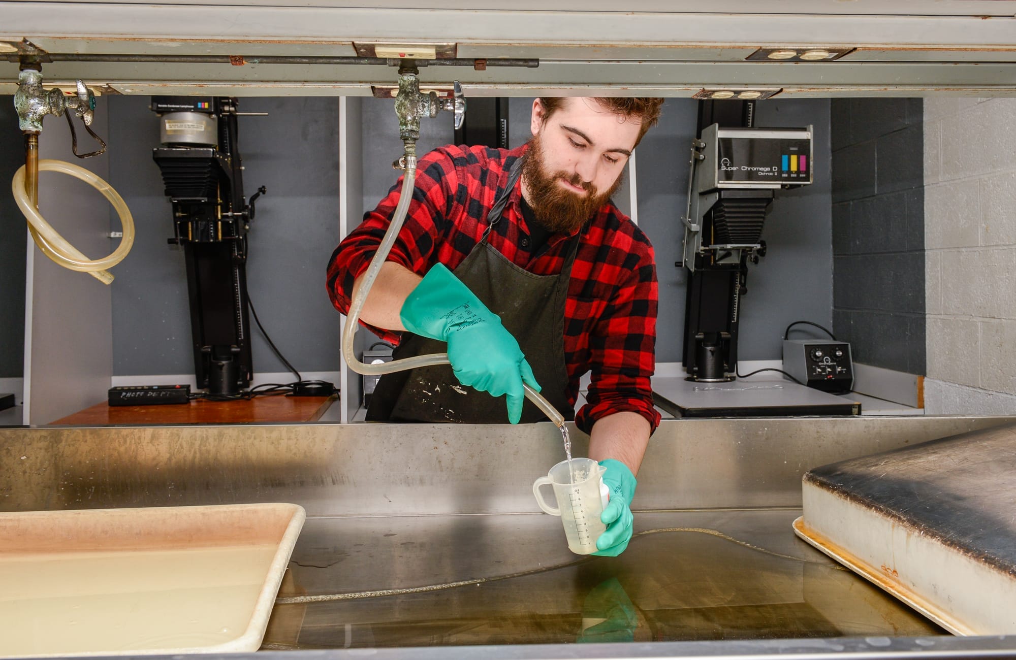 A man with a beard, wearing a red plaid shirt and green gloves, is working in a darkroom. In this self-service setup, hes measuring liquid into a jug on a workbench, surrounded by photo enlargers and trays in the background.