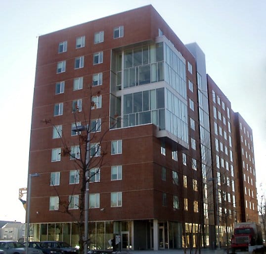 A modern, multi-story brick apartment building with large glass sections and numerous windows. The structure stands tall against a clear sky, with trees and a street lamp visible in the foreground.