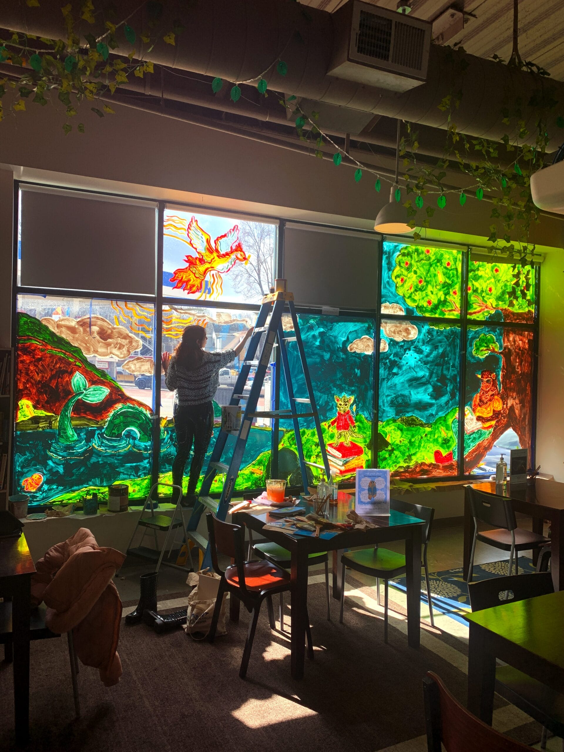A person stands on a ladder painting vibrant, colorful fantasy scenes on large café windows as part of a PartnershipWORKS project. Sunlight streams through the artwork, illuminating the cozy, art-filled room.