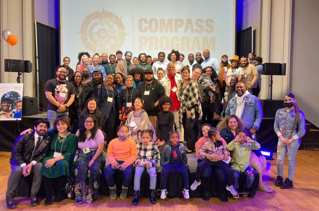A diverse group, including joyful adults and children, gathers on stage before a screen that proudly displays Compass Program. The atmosphere is celebratory, with smiles all around as they hold children and pose for the perfect SEO keywords-enhanced photo.