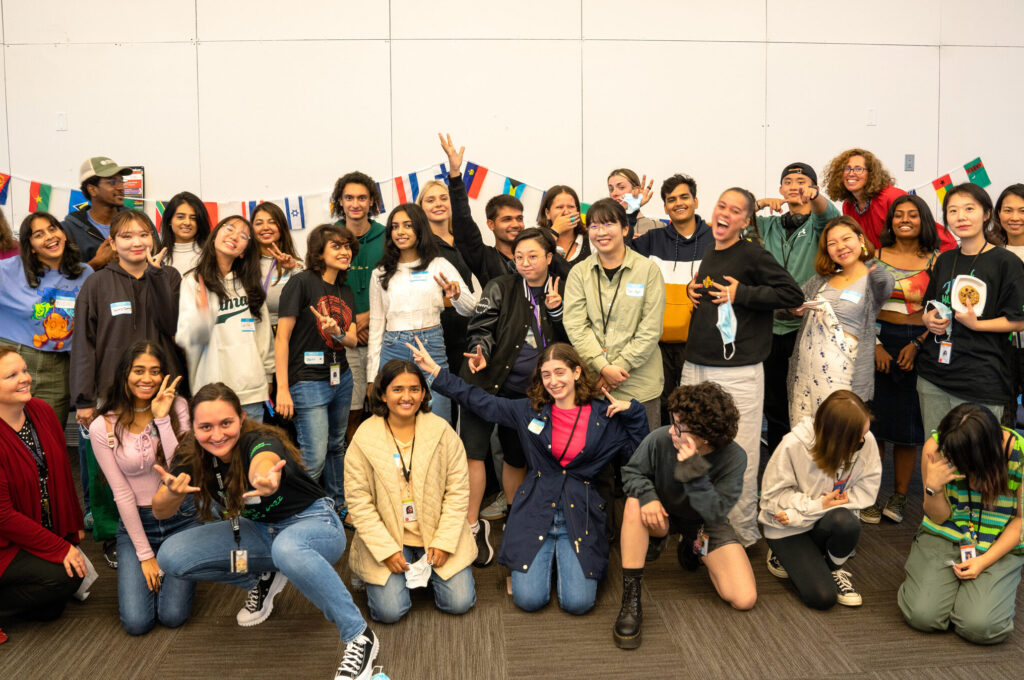A diverse group of students in vibrant clothing pose and smile in a room adorned with international flags. Some individuals are gesturing playfully, creating a lively and supportive atmosphere against the backdrop of a plain wall.