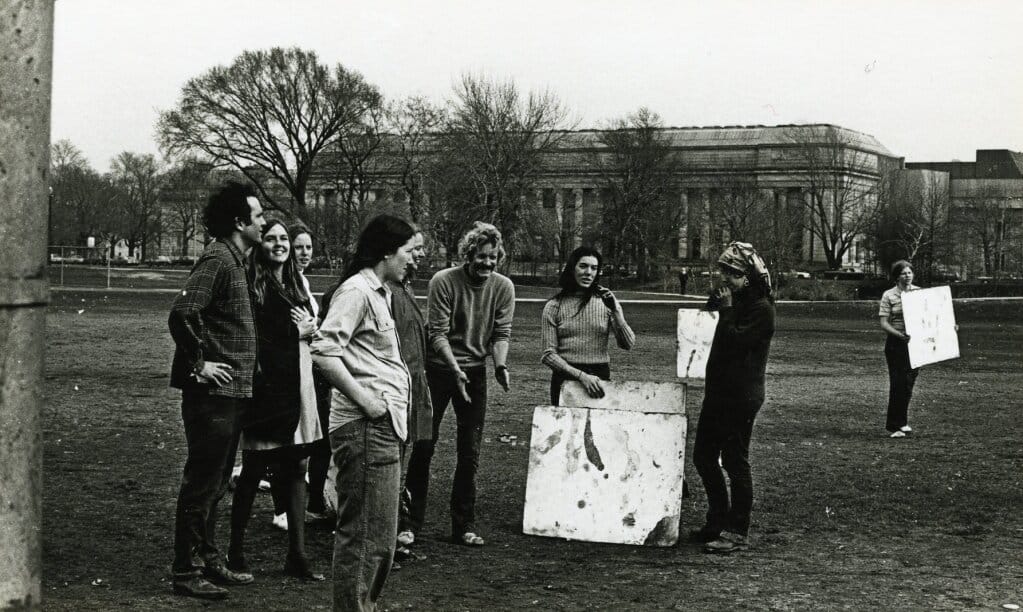 A group of young adults casually gather outdoors on a grassy field with bare trees and a large building in the background. Some are observing painted canvases on the ground. The scene has a candid, social atmosphere.