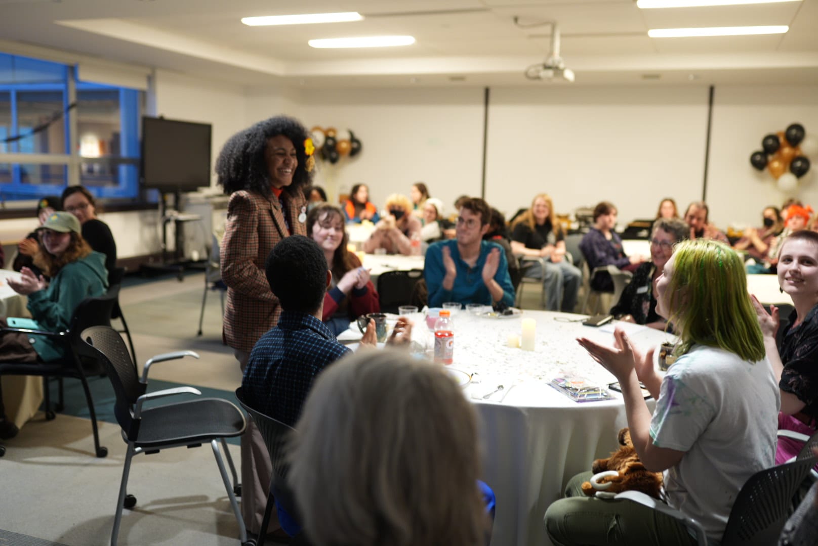 A woman stands smiling as people seated at round tables applaud her in a well-lit room. As a student leader, she beams with pride. The audience is engaged and cheerful, with drinks, decor, and balloons adding to the festive atmosphere.