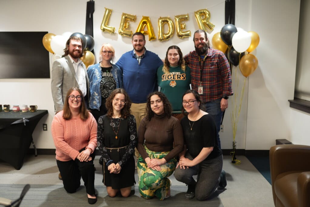 A group of student leaders poses in a room with gold and black balloons. A balloon arrangement above spells LEADER. They are casually dressed, smiling, and some are seated while others stand behind them.