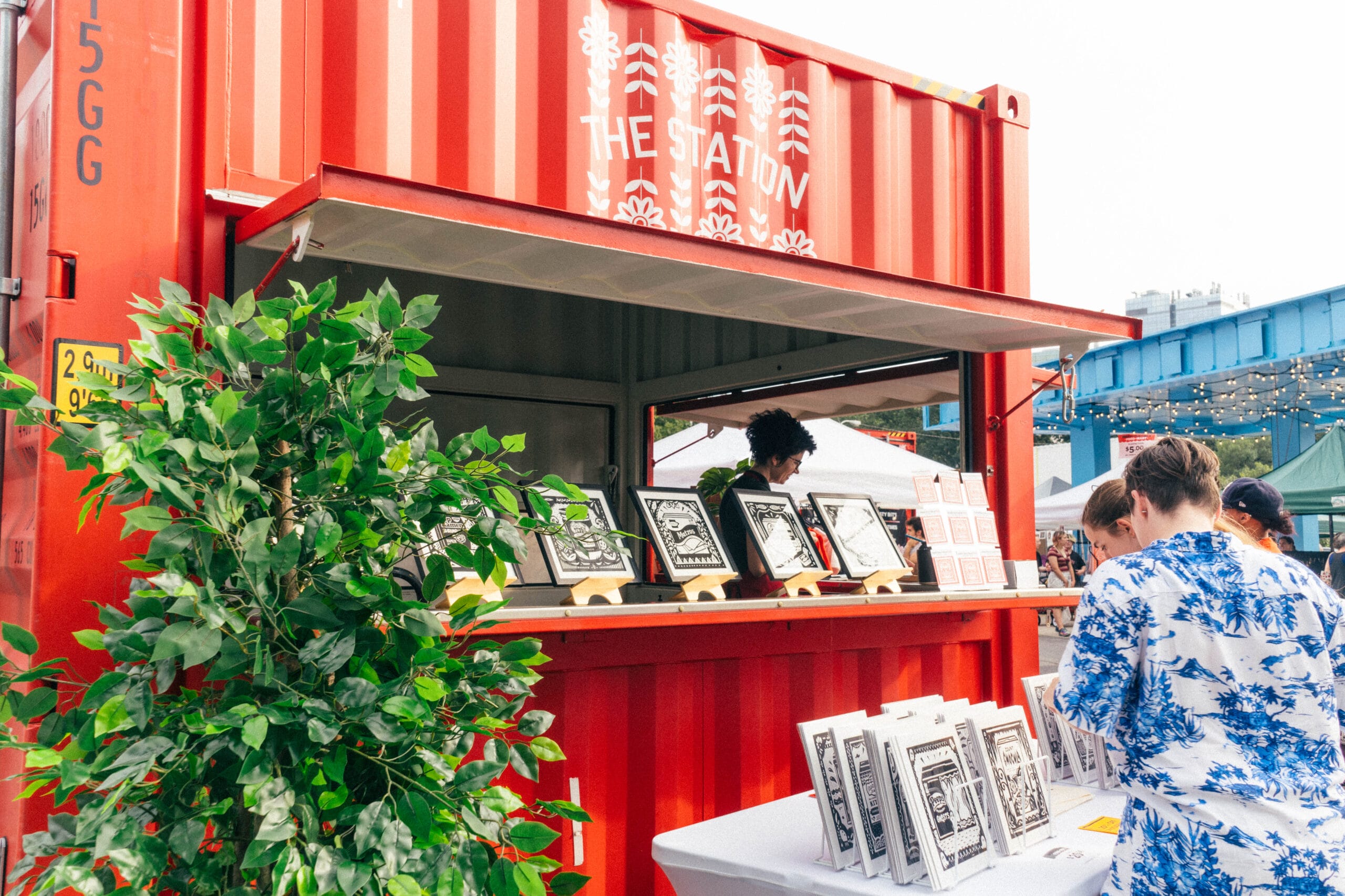 A red shipping crate showcases an artists' work at an outdoor art market.