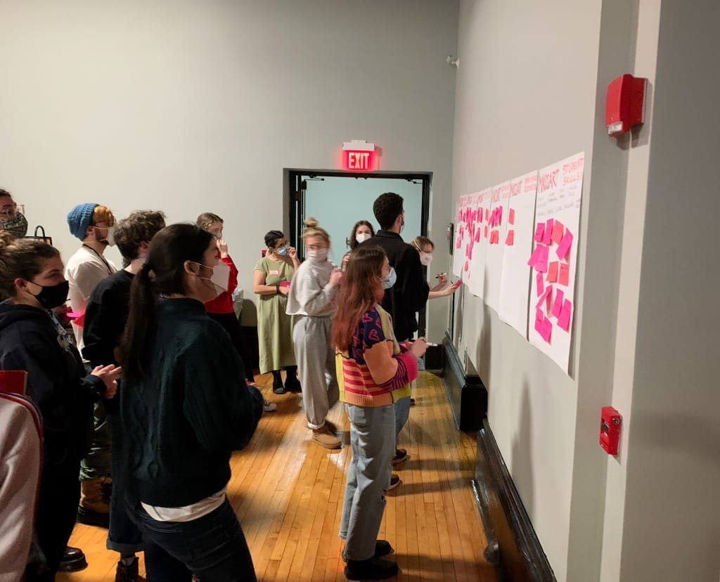 A class of students in a gallery setting looking at a wall of post-it notes