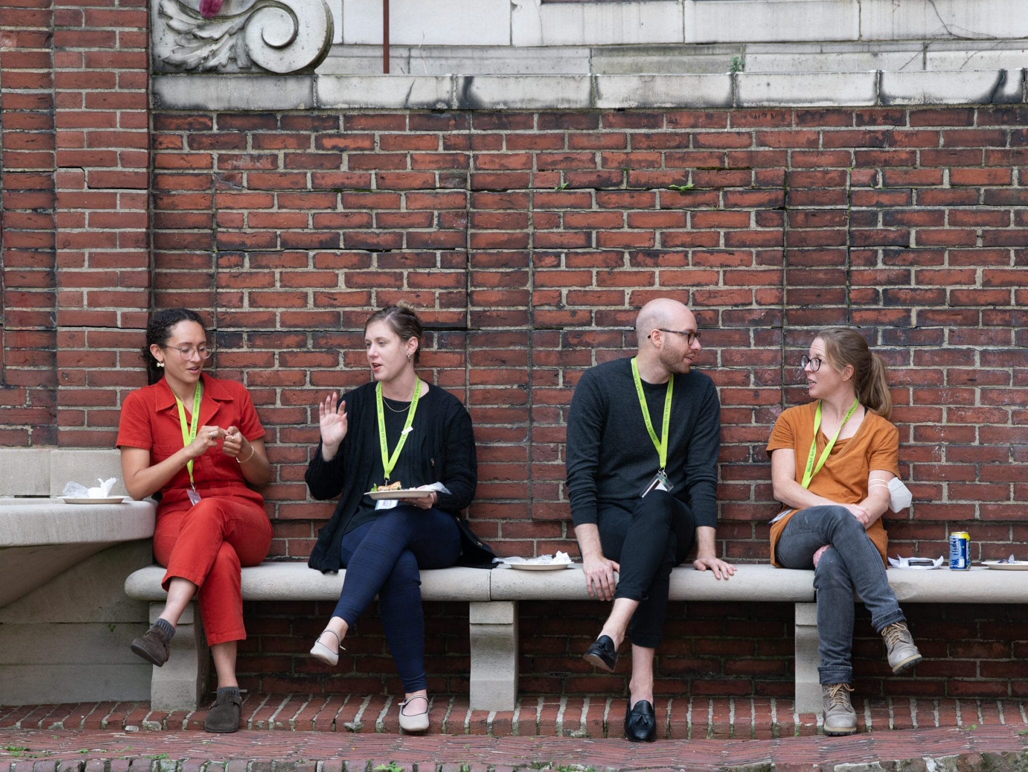 Four people sit on a bench against a brick wall, wearing conference lanyards. Three are engaged in conversation about work opportunities, while one holds a plate of food. They appear relaxed, with casual attire, in an outdoor setting at MassArt.