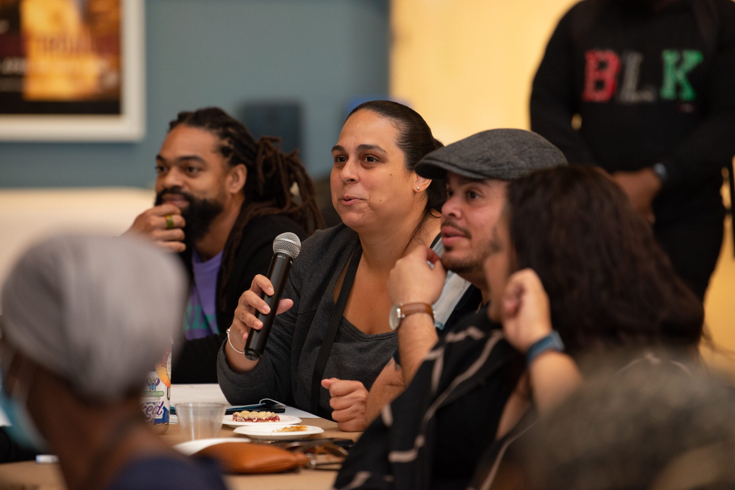 A group of people sitting at a table attentively listening. One person holds a microphone, appearing to speak or ask a question. Plates and cups are on the table, suggesting a discussion during a meal or event.