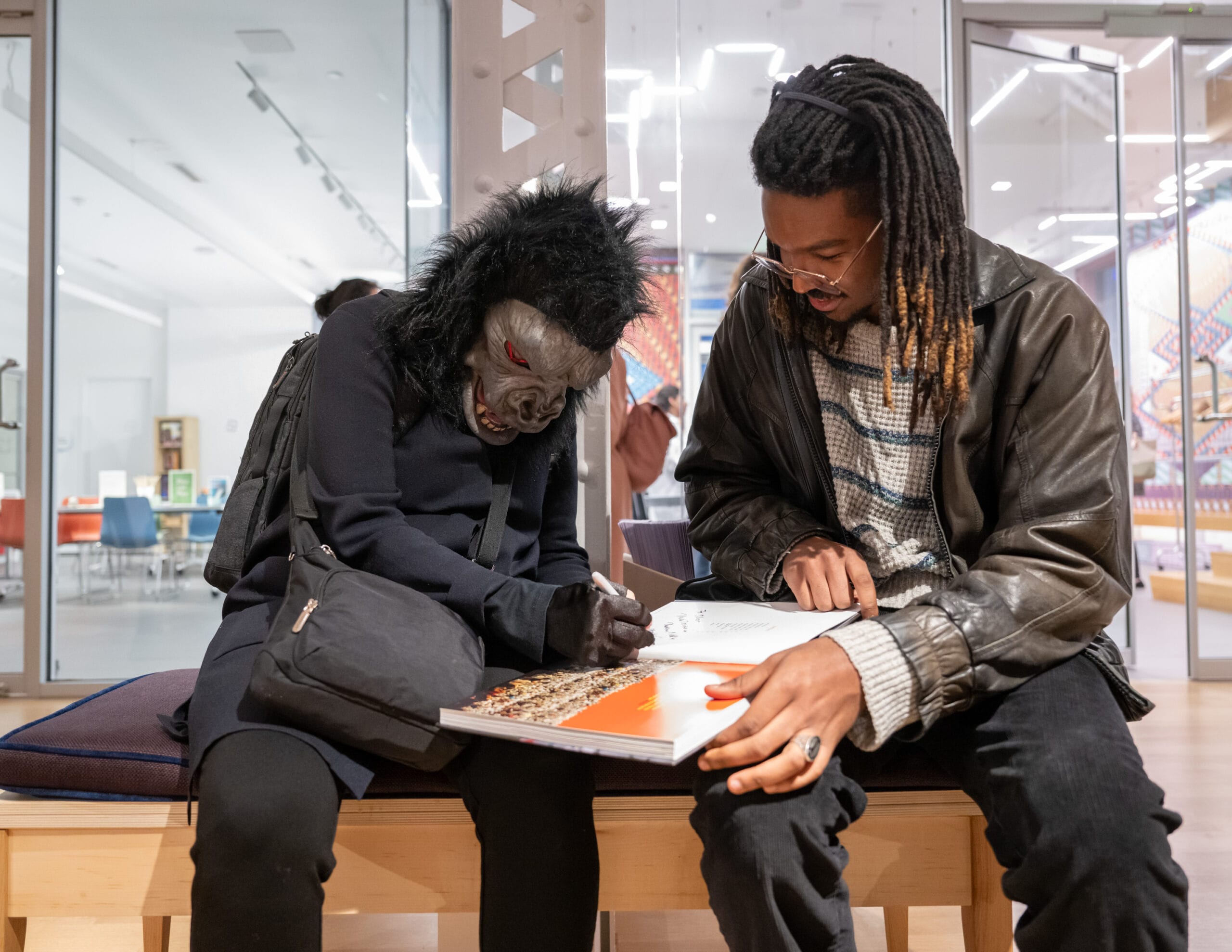 A person in a guerilla costume sits and signs a book held by a visitor to the MassArt Art Museum.