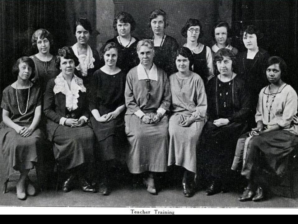 A black and white photo of thirteen women sitting and standing, posing formally. They are all dressed in modest early 20th-century clothing, with the caption Teacher Training below them. The background appears dark and plain.