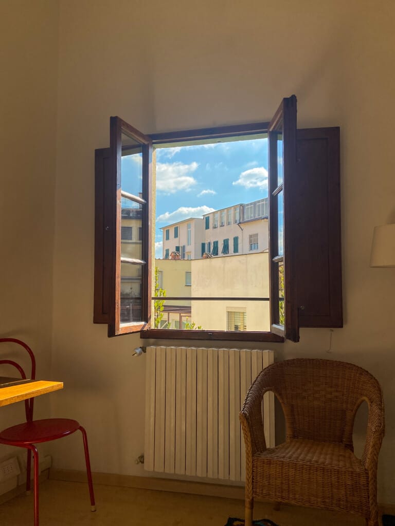 Sunlit room with an open window revealing a view of neighboring buildings against a blue sky with scattered clouds, perfect for contemplating your next creative project when you study abroad at MassArt. Inside, find a wicker chair, red chair, and part of a wooden desk by the radiator.