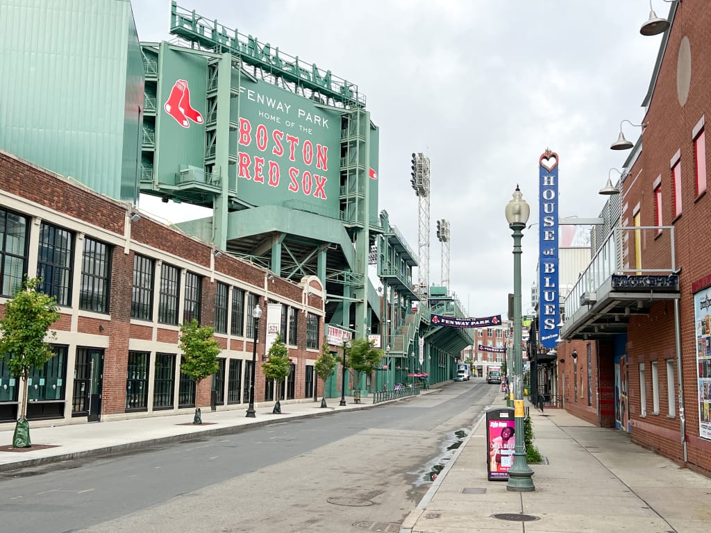 Street view of Fenway Park, home of the Boston Red Sox, with a large green sign and baseball fields visible. The street is empty, flanked by brick buildings. A House of Blues sign hints at the vibrant performance venues nearby under an overcast sky.