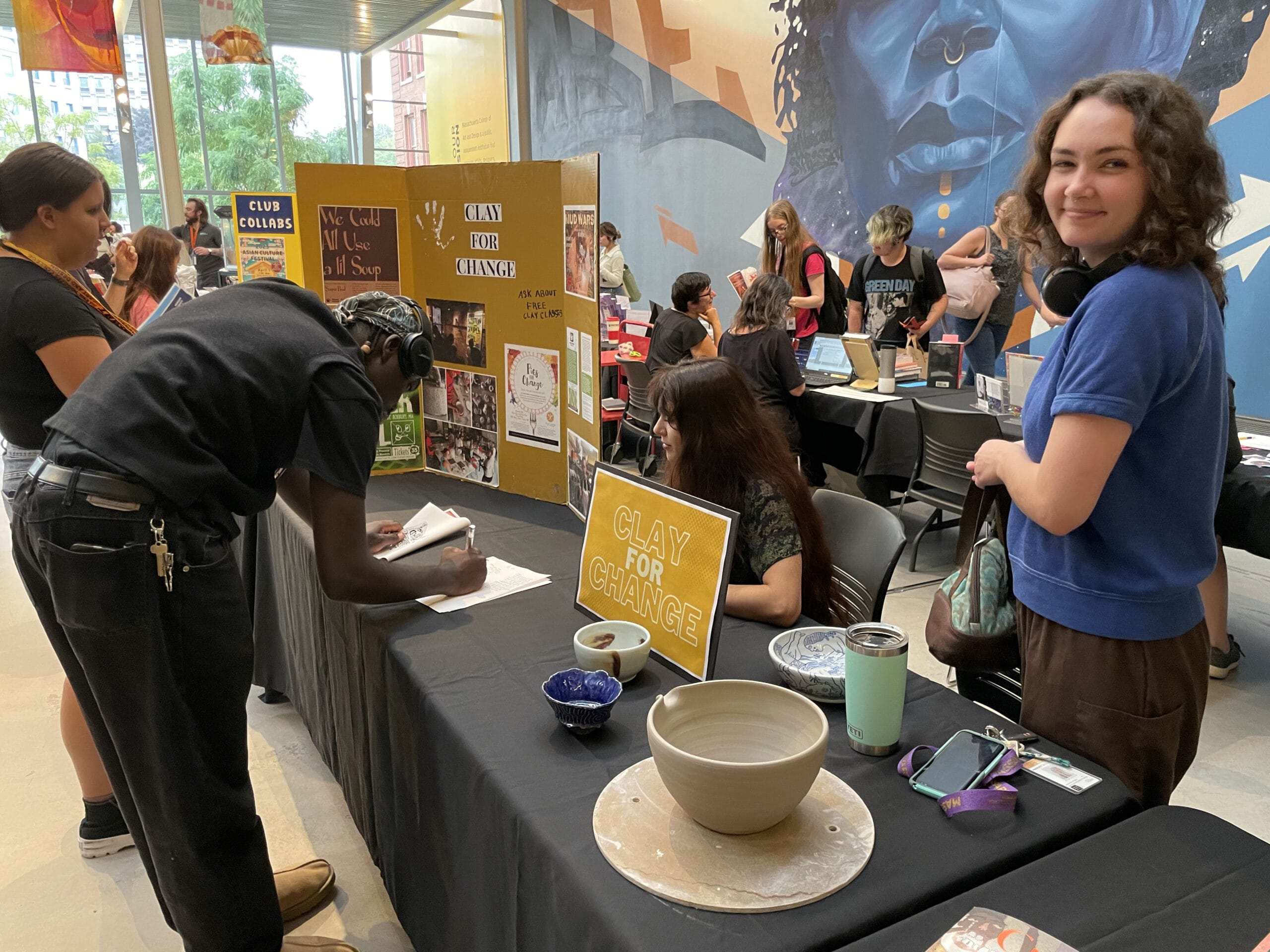 People attending a campus club fair inside a building. A person signs up at the Clay for Change table, which has pottery items displayed. Others are engaging with different booths. The atmosphere is lively and social.