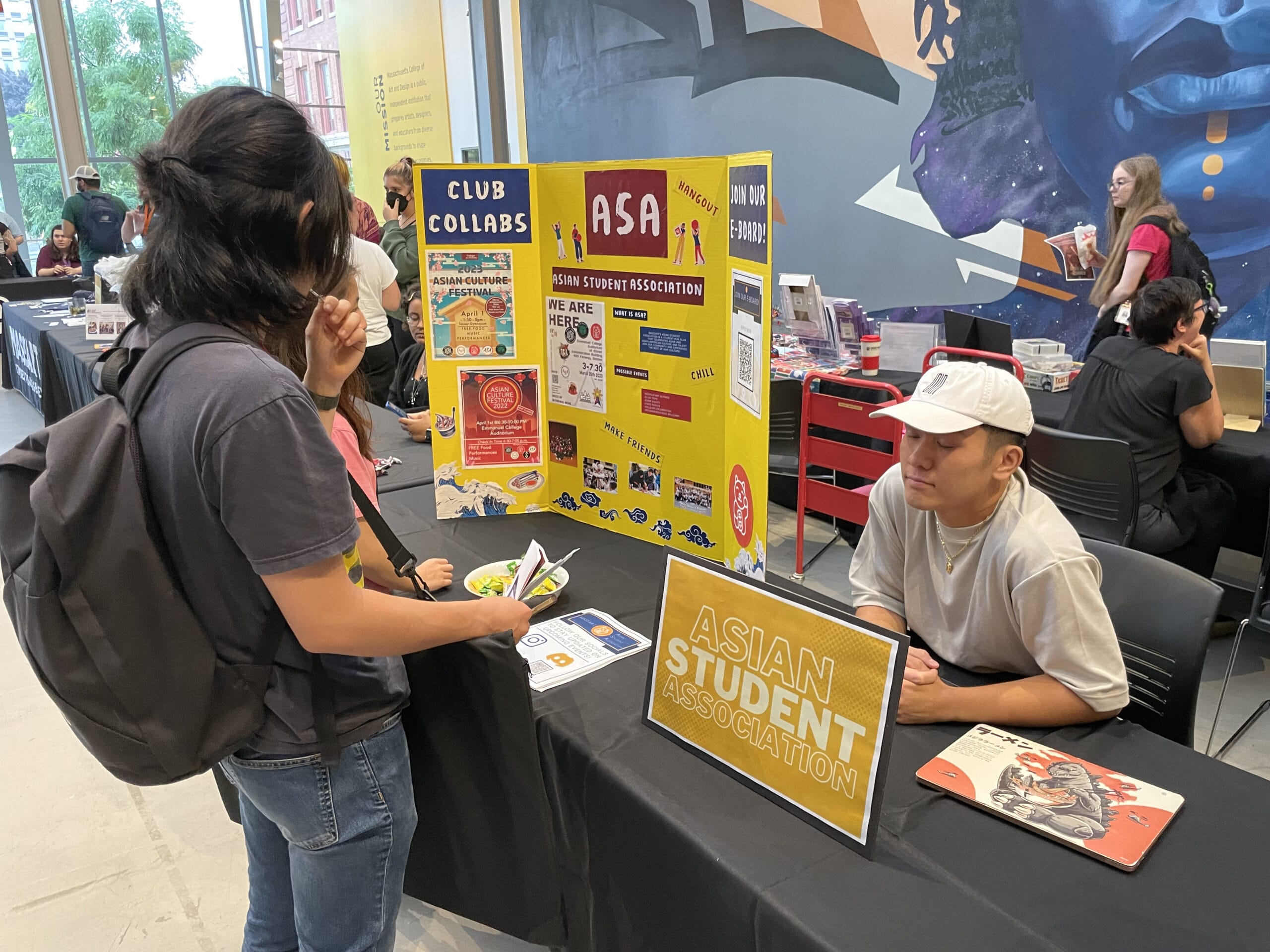 Two people engage at an Asian Student Association booth during a club fair. The booth displays colorful posters and pamphlets. A person with long hair holds a brochure, while another in a white cap sits behind the table with a laptop.