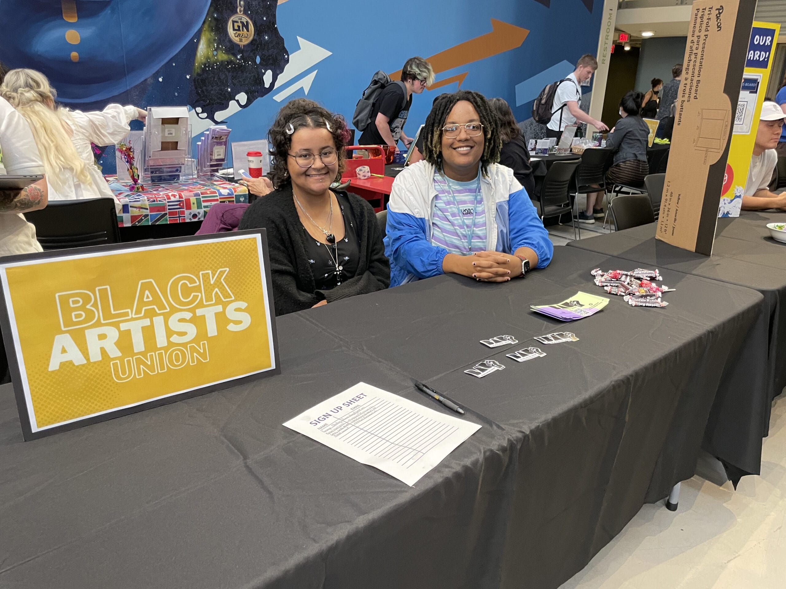 Two people seated at a table with a Black Artists Union sign. The table has flyers, a sign-up sheet, and other materials. People are visible in the background at an event with colorful walls.