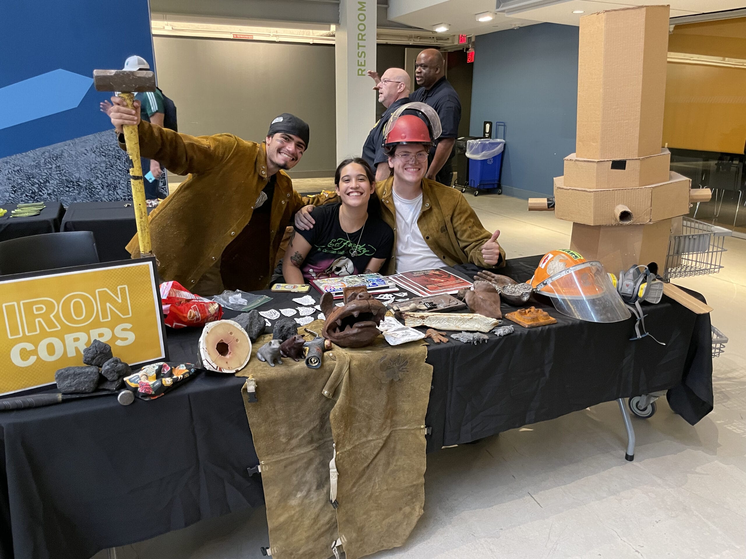 Three people smile at a booth with an Iron Corps sign, promoting student organizations. The table displays items like a hard hat, brochures, and rocks. One person holds a sledgehammer. Nearby, cardboard boxes are stacked high, with two more people in the background.