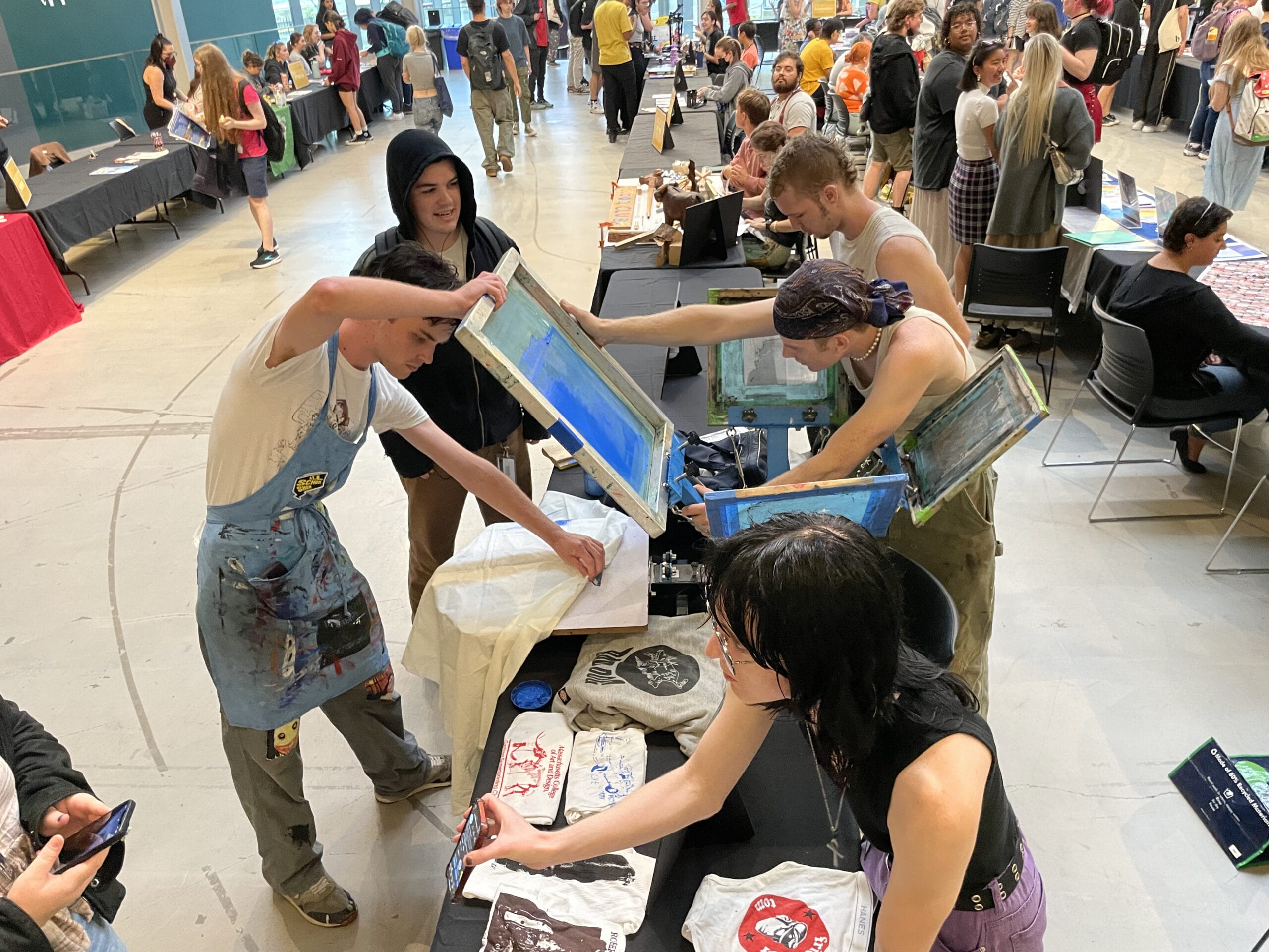 During a lively campus art event, students from various clubs are screen printing designs on fabric at a bustling table. Others gather around, eager to participate or admire the work, while art pieces displayed nearby contribute to the creative atmosphere.
