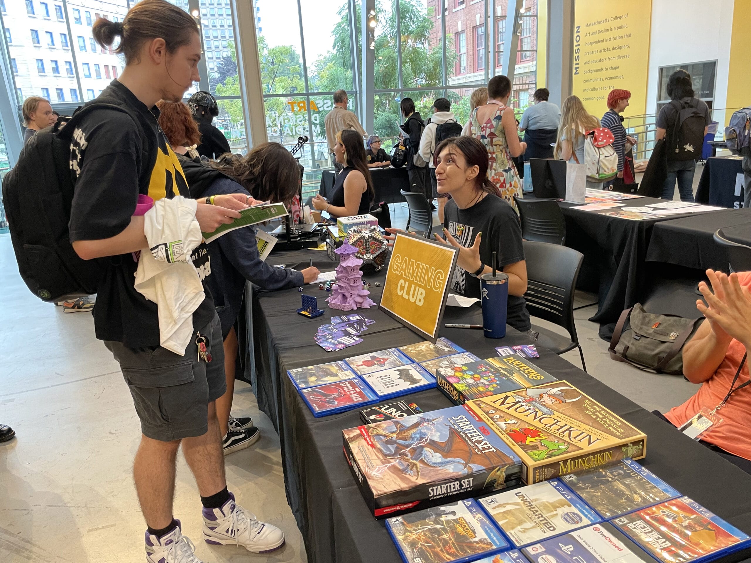 People are gathered around a table at a campus activities gaming club event. A person standing holds a board game while another seated person talks to them. Various games, including Munchkin and a Starter Set, are displayed as other students browse in the background.
