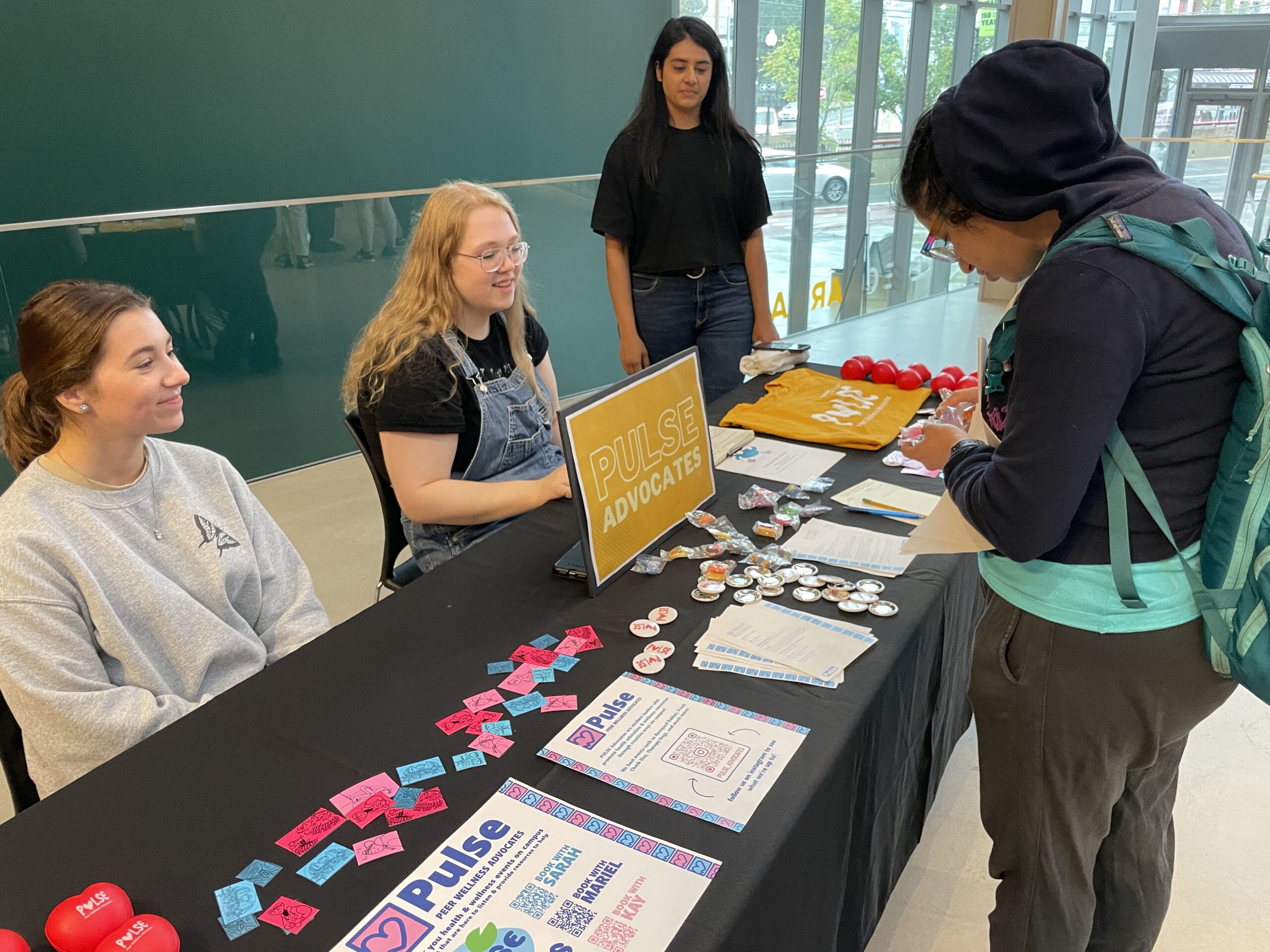 A vibrant scene unfolds as people gather around a table marked by a Pulse Advocates sign, featuring pamphlets, buttons, and heart-shaped stress balls. In this bustling hallway setting, a person with a backpack diligently fills out a form while student organization members engage nearby.