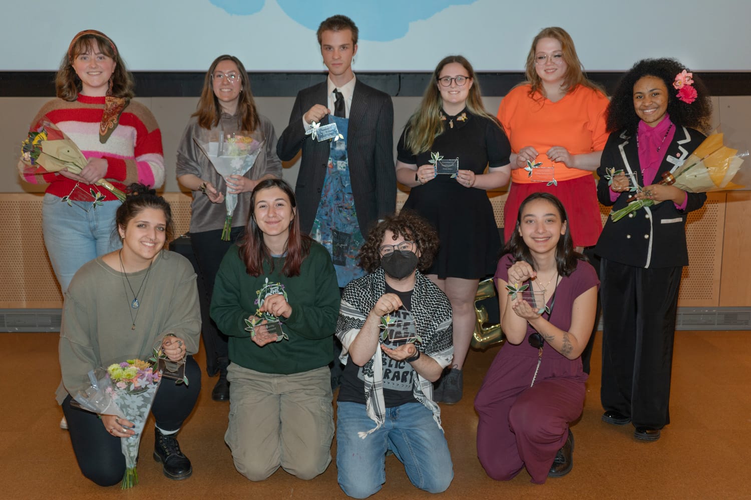A group of ten people pose in two rows, embodying student leadership. The back row holds bouquets and small objects, dressed in a mix of casual and formal attire. Kneeling in the front, some hold flowers; one sports a mask. A large screen looms behind them, signaling leadership development.