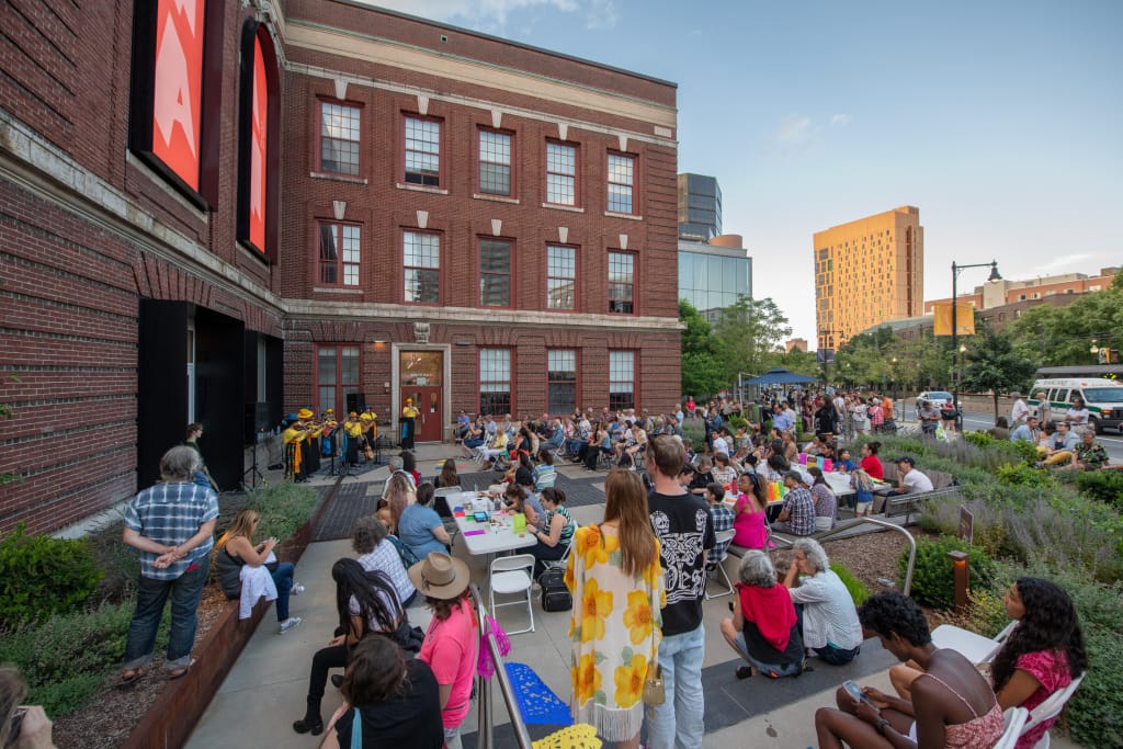 A large crowd gathers in an outdoor space beside a brick building for a summer performance. A group of musicians plays on a small stage as attendees, seated at tables and steps, enjoy the event. The sky is clear, and several housing structures are visible in the background.